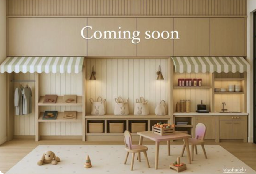Empty children's playroom with a small table, two chairs, shelves with baskets, a stuffed toy, and a nursery set. The wall has a ‘Coming soon’ sign.