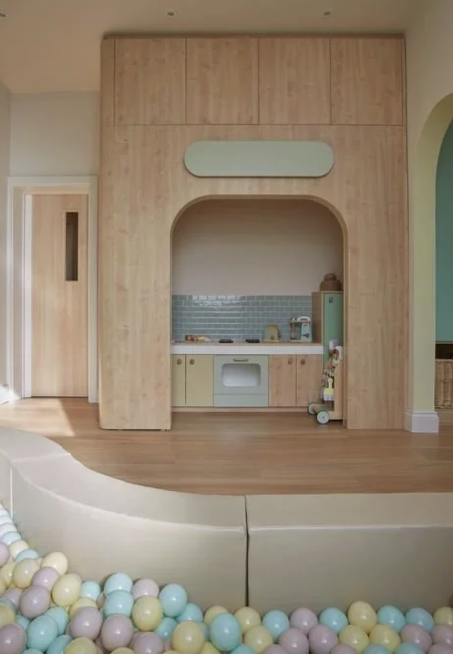 Children's play area with a small kitchenette built into wooden wall, pastel-colored ball pit in foreground.