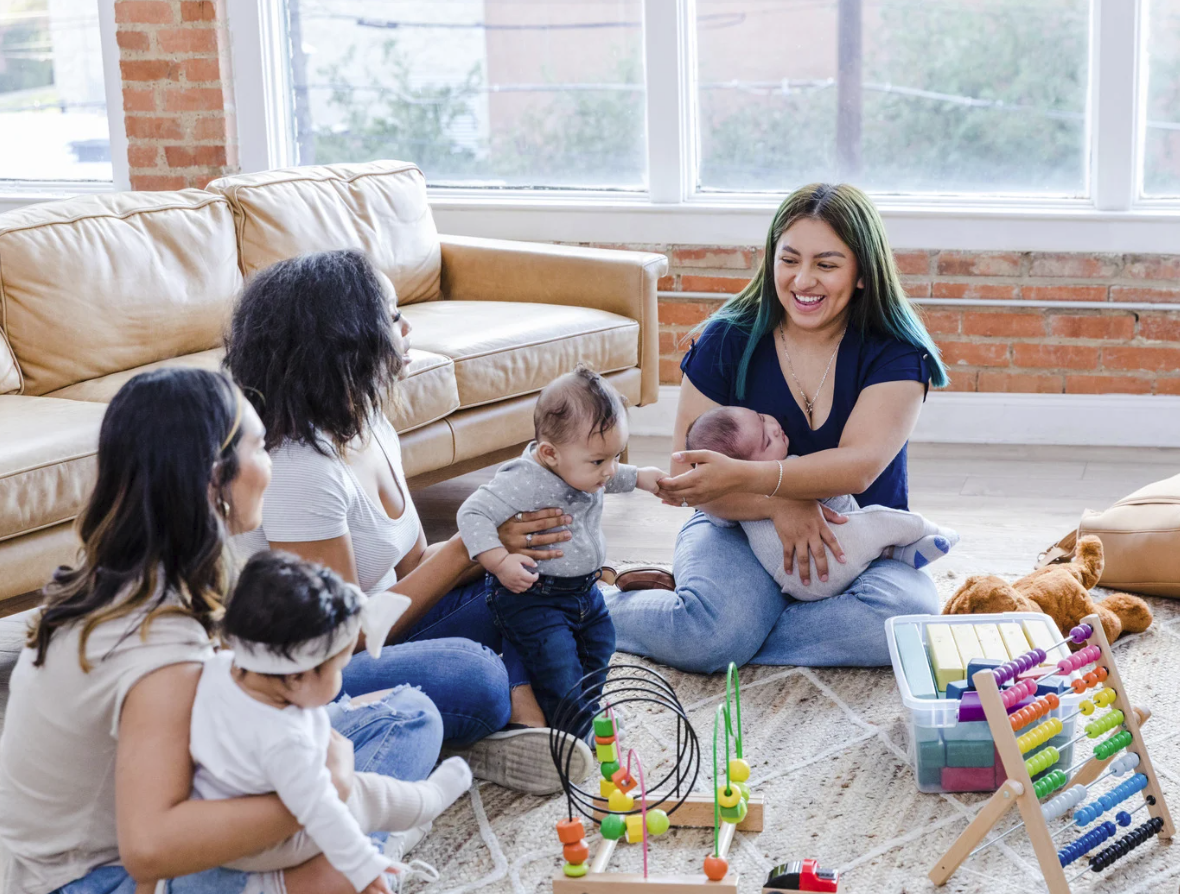 A woman with green hair sits on the floor holding a newborn, smiling at a group of diverse women and children sitting around her in a living room with a brick wall and large windows.