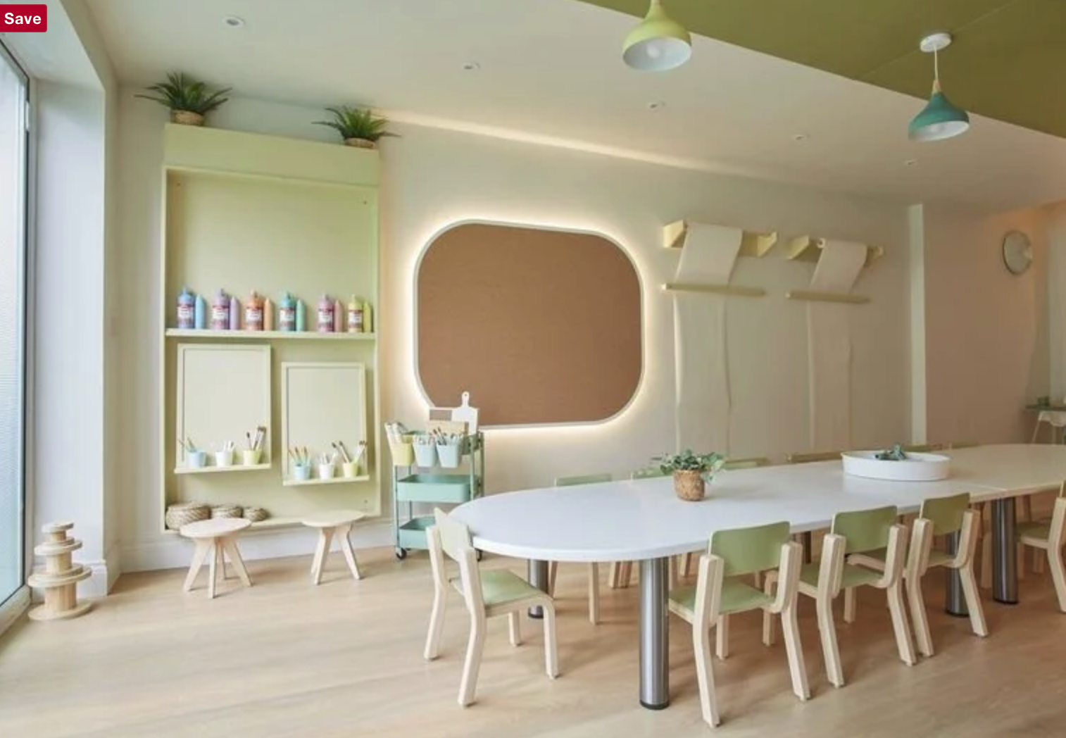 Bright, modern room with pastel green and beige decor, large rounded mirror with backlighting, a long white table with green chairs, shelves with art supplies, potted plants, and natural light from large windows.