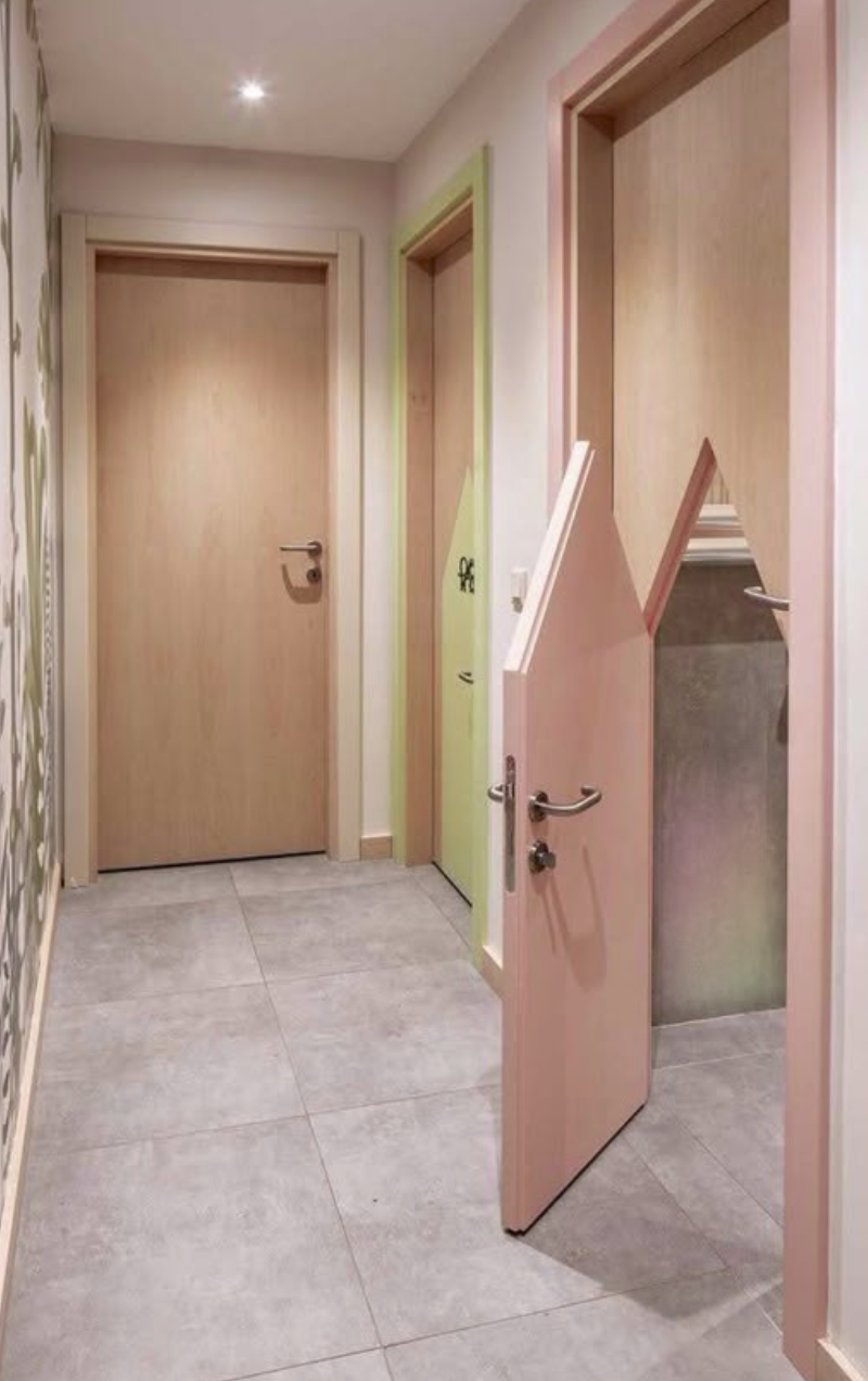 A hallway with three wooden doors, one pink, one pale green, and one light wooden, with a colorful wall on the left and tiled floor.