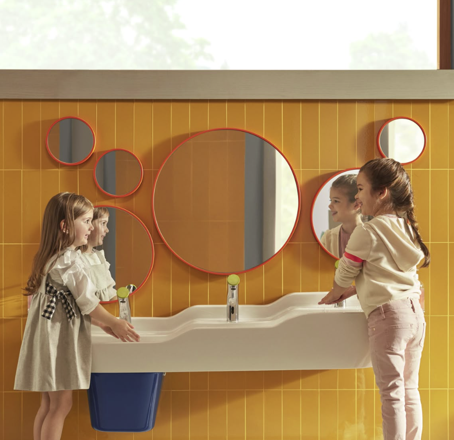 Two young girls are looking at and touching a shared communal bathroom sink with three mirrors on a yellow tiled wall. One girl is wearing a beige dress with black bow accents, and the other is dressed in a white hoodie and pink pants. The large cent