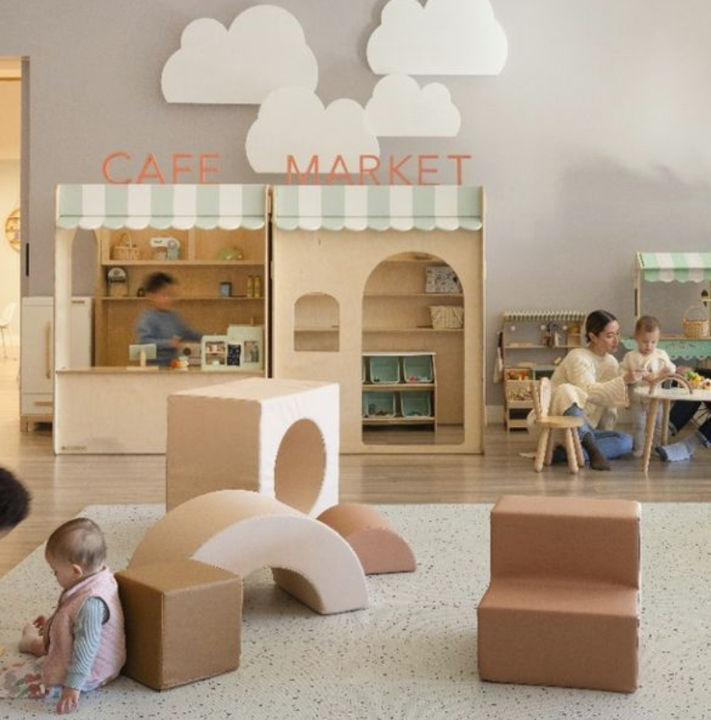 Children playing in a modern indoor play area with pastel-colored foam blocks, a wooden play market stand, and wall decorations of clouds.