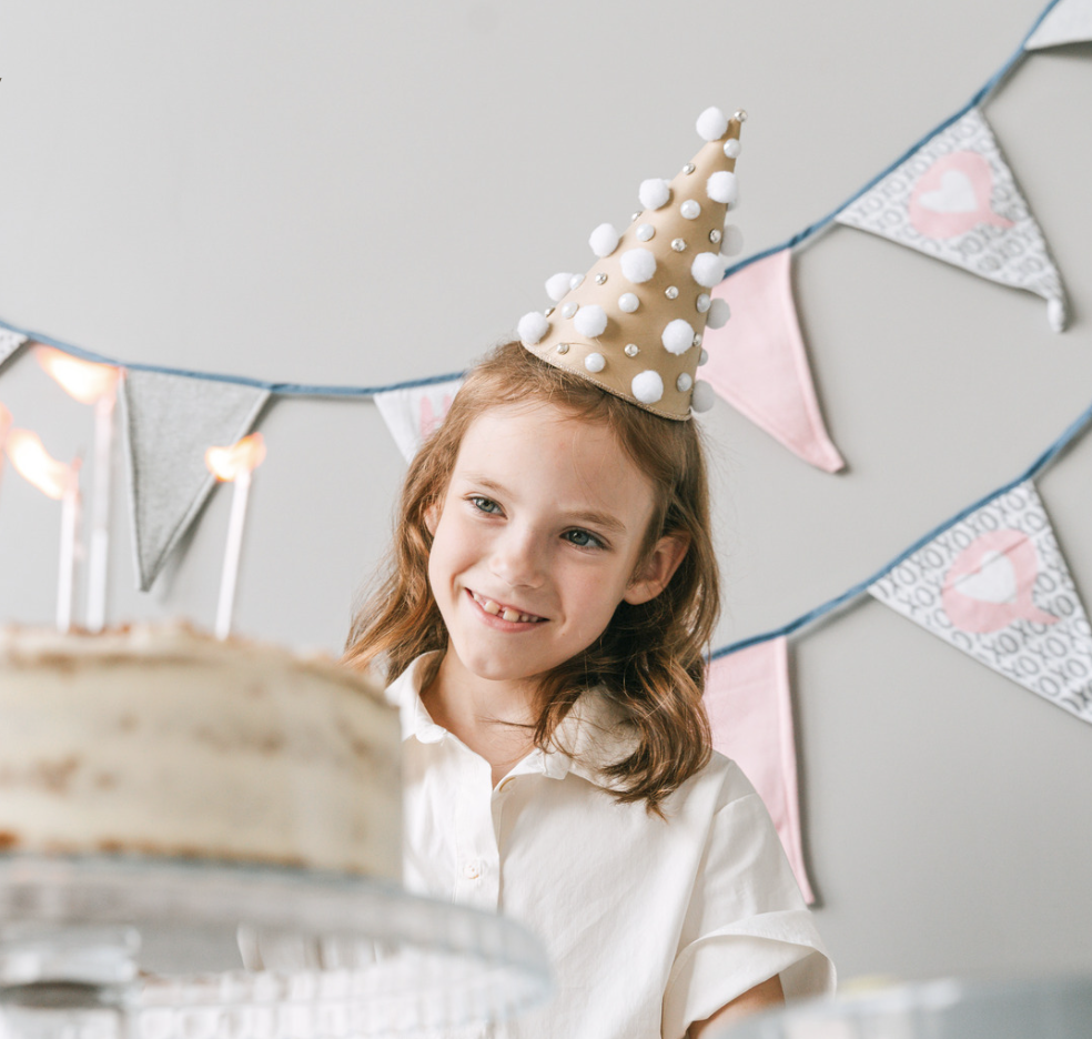 A girl wearing a party hat with pom-poms and pearls, celebrating a birthday with a cake in front of her, and birthday decorations in the background.