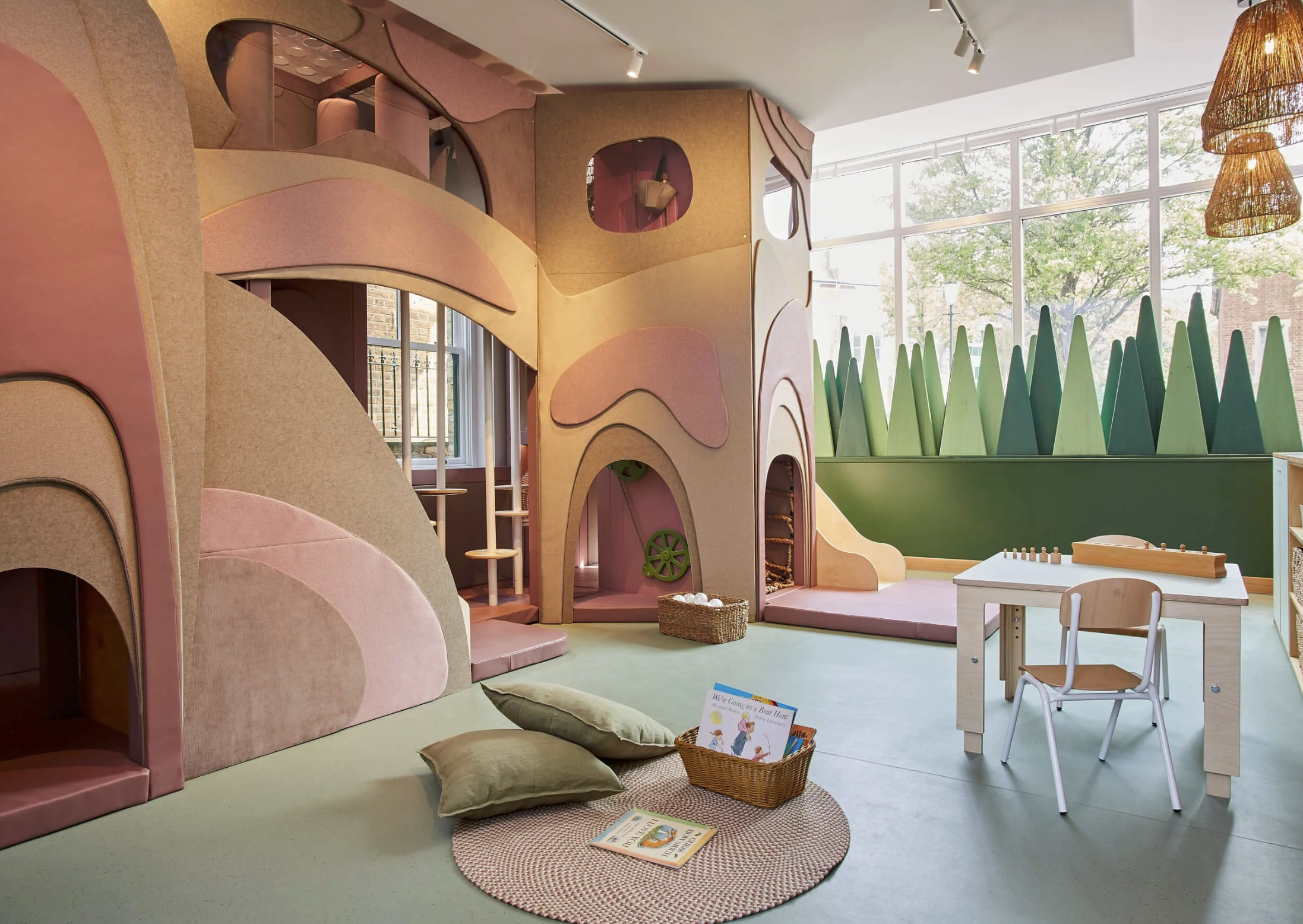 Indoor children's play area with pastel-colored foam climbing structures, a slide, a small table with chess, a chair, and a basket of books and pillows on a circular rug. Large window with greenery outside.