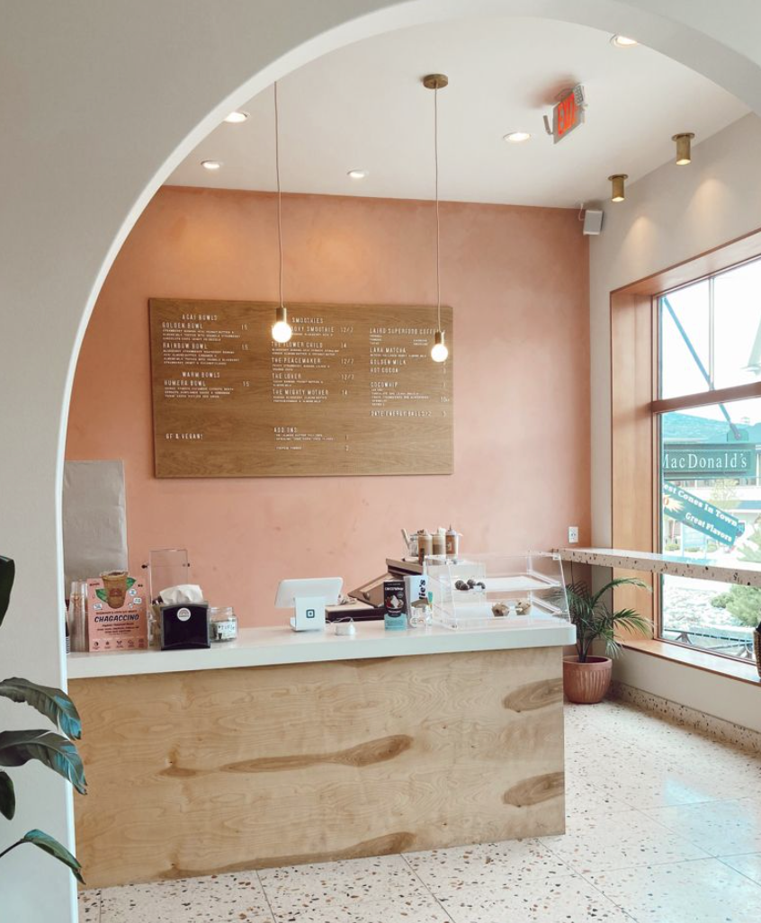 A modern, minimalist coffee shop counter with a light wood finish, a large menu board on a peach-colored wall, and a window showing a glimpse of a MacDonald's outside. Potted plants are near the window.