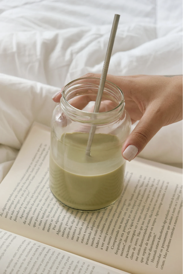 Hand holding a glass jar with a straw, filled with a green smoothie, next to an open book on a bed with white sheets.