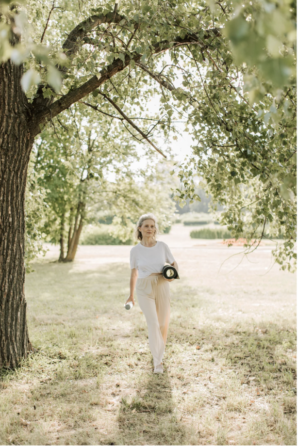 A woman walking outdoors in a park-like setting under a large leafy tree, holding a small bottle and a hat, with sunlight filtering through the leaves and a grassy pathway.