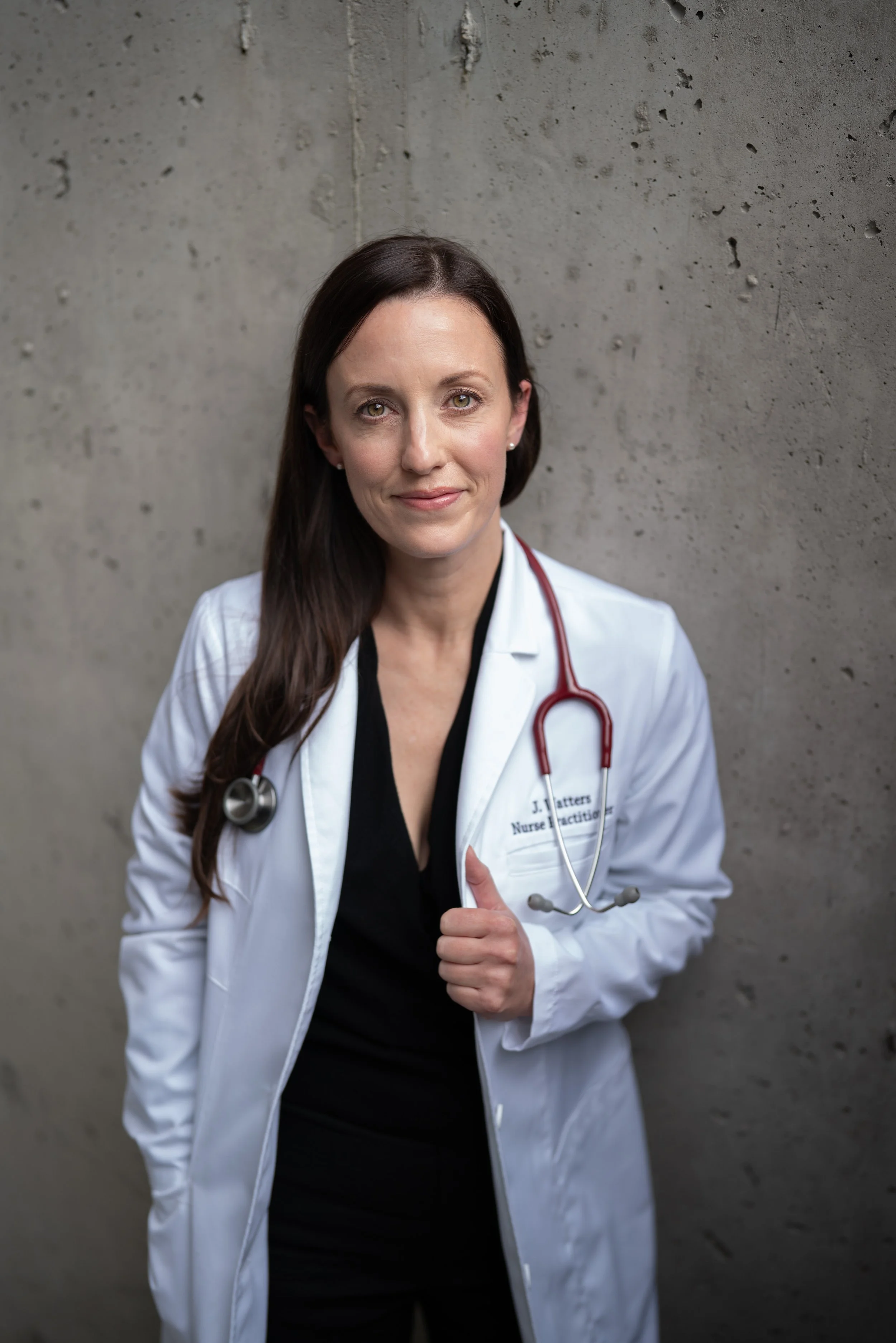 A picture of Jennifer Watters, MN NP(A) MSCP Certified Menopause Practitioner with long brown hair wearing a white coat and stethoscope giving a thumbs-up against a concrete wall.