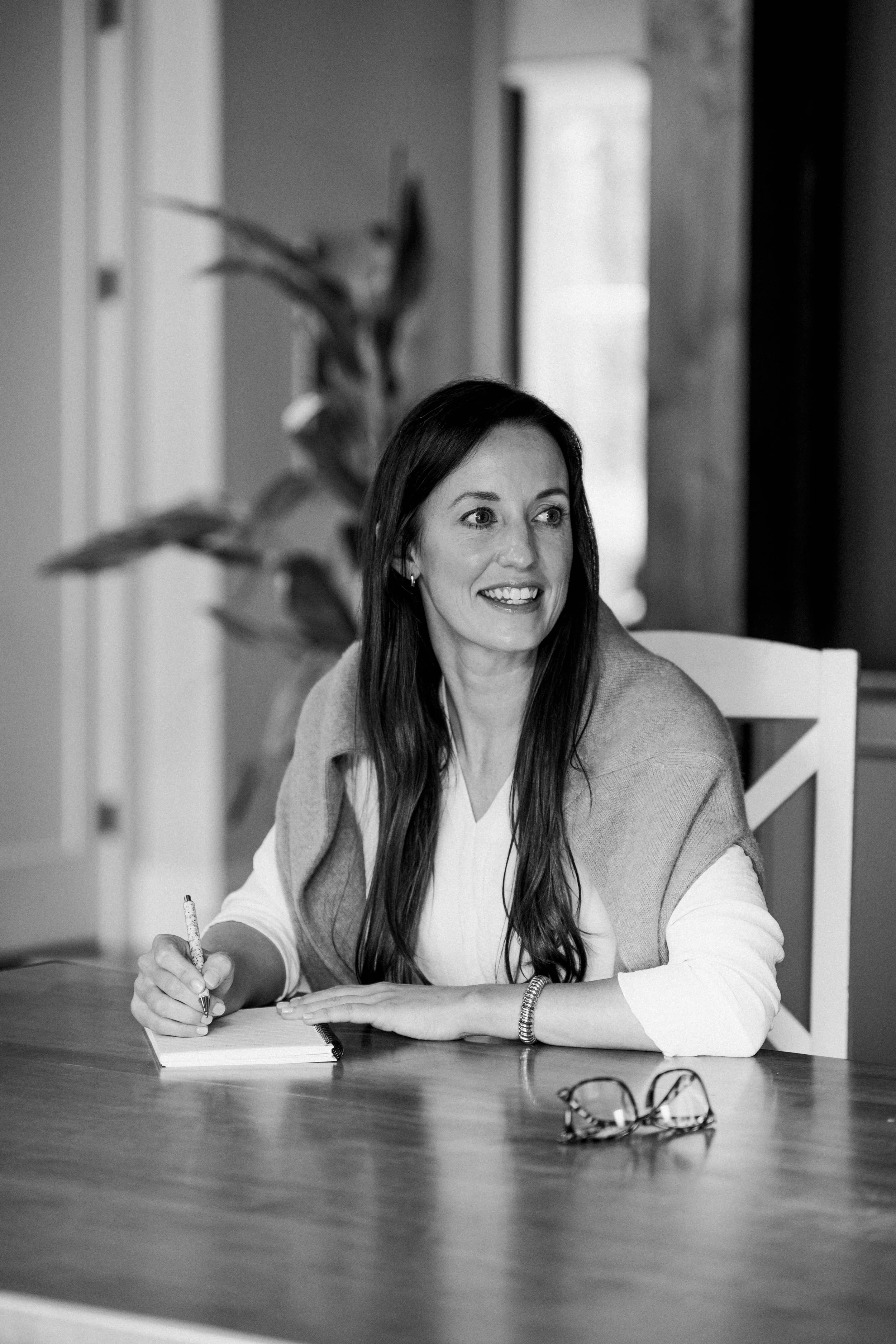 A woman with long dark hair sitting at a wooden table, smiling, holding a pen, with glasses on the table, in a room with a window and a plant in the background.