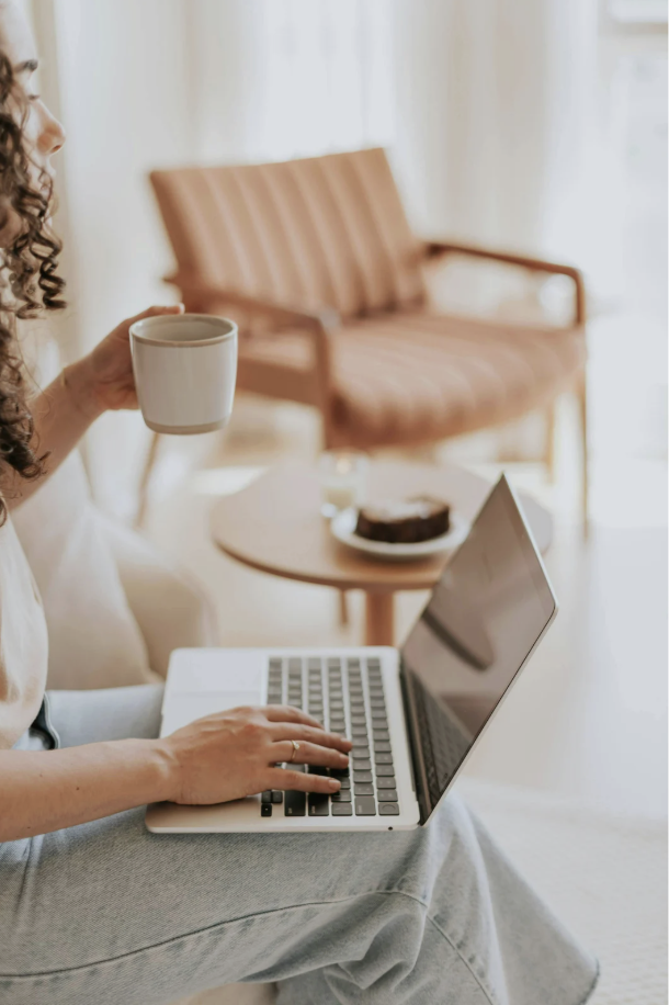 Person working on a laptop, holding a coffee mug, with a cake on a table behind.