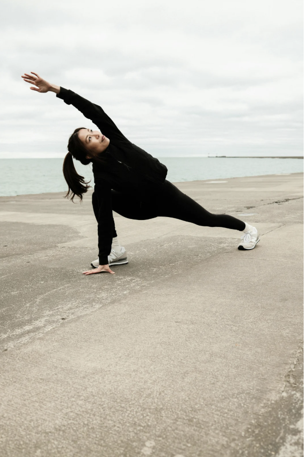 A woman in black athletic wear performs a side plank yoga pose on a concrete surface near the water with cloudy sky overhead.