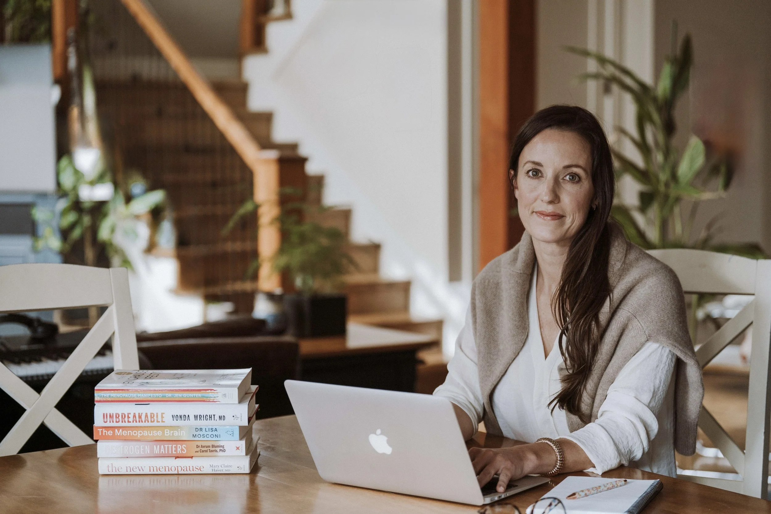 A picture of Jennifer Watters, MN NP(A) MSCP Certified Menopause Practitioner sitting at a wooden table with a stack of medical, health, wellness perimenopause  menopause books, a laptop, and a notebook. Background is a staircase, plants, cozy home.