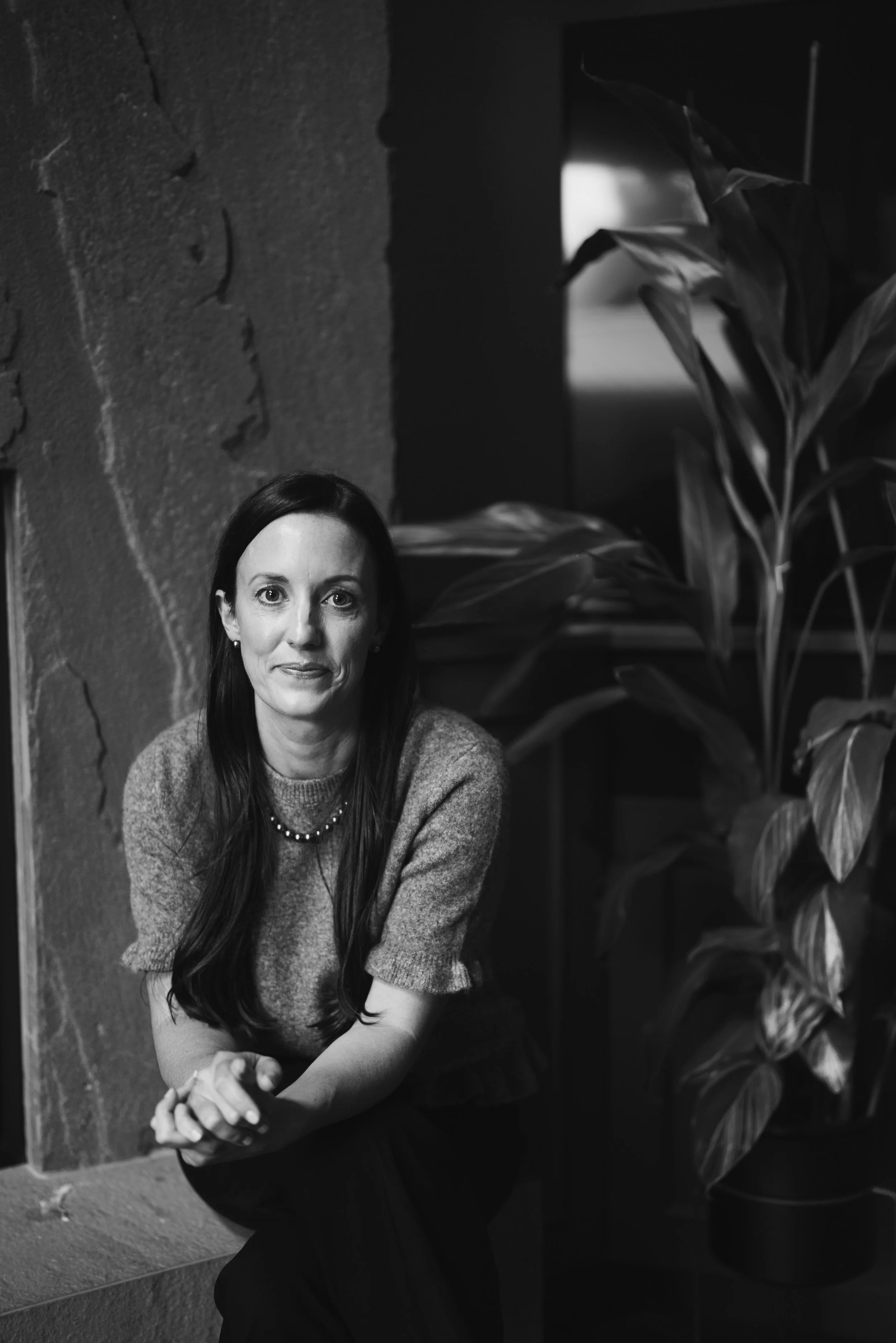A black and white photo of Jennifer Watters, MN NP(A) MSCP Certified Menopause Practitioner with long dark hair and a sweater, sitting beside a textured wall and a large leafy plant. RoseWell Health