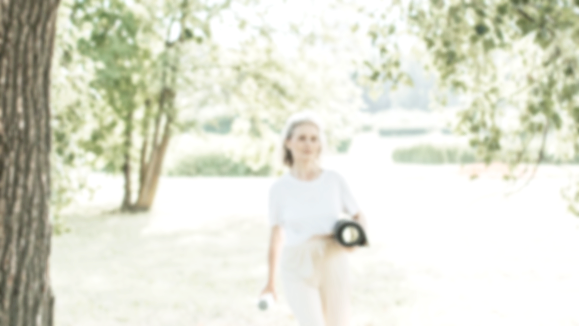 A woman walking in a park or outdoor area during daytime, holding an object in her right hand, with trees and greenery in the background.