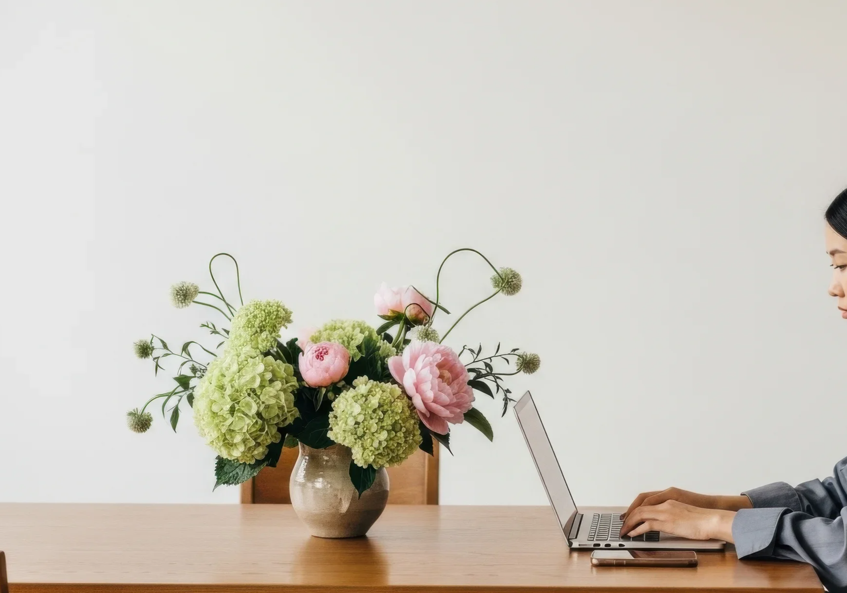 Person in gray blazer working on a silver laptop at a wooden table with a large bouquet of pastel pink and green flowers in a beige ceramic vase against a plain white wall.