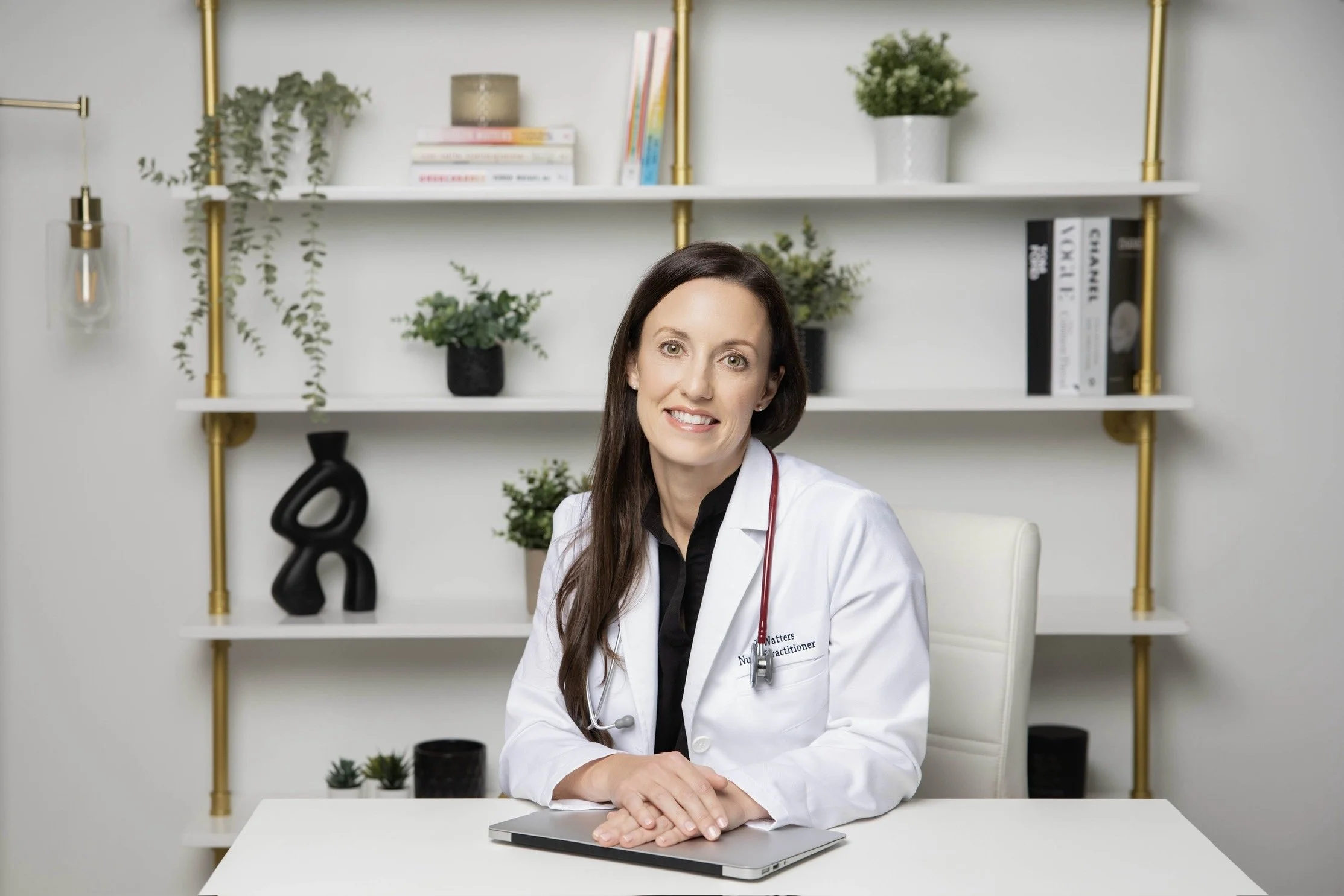 Jennifer Watters, nurse practitioner, RoseWell Health, sitting at a white desk in an office, warm smile. She is wearing a white coat and stethoscope. Behind her is a white shelf, plants, medical books. Virtual consultation visit