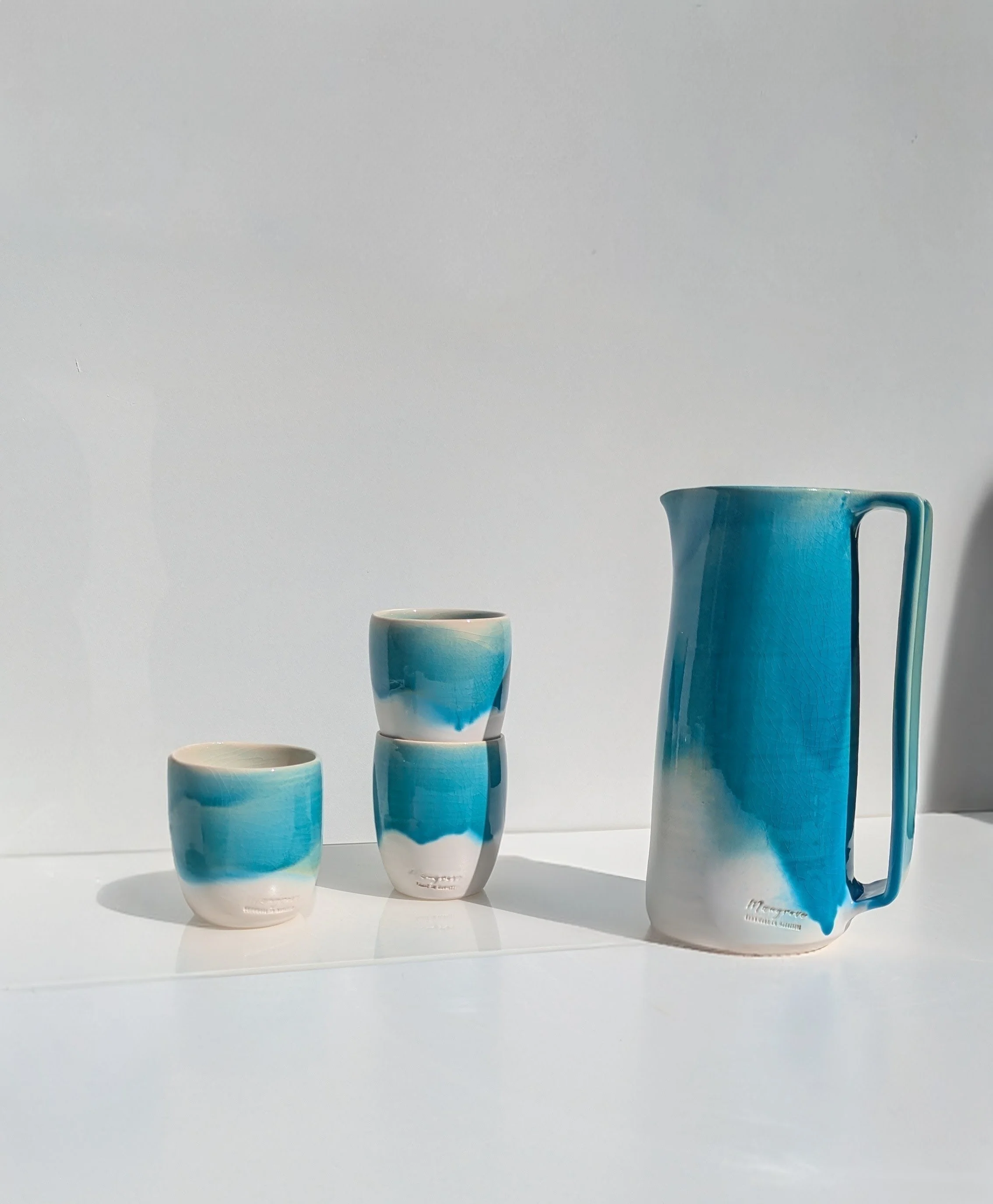Four ceramic cups and a pitcher with blue and white glaze, arranged on a white surface against a plain background.