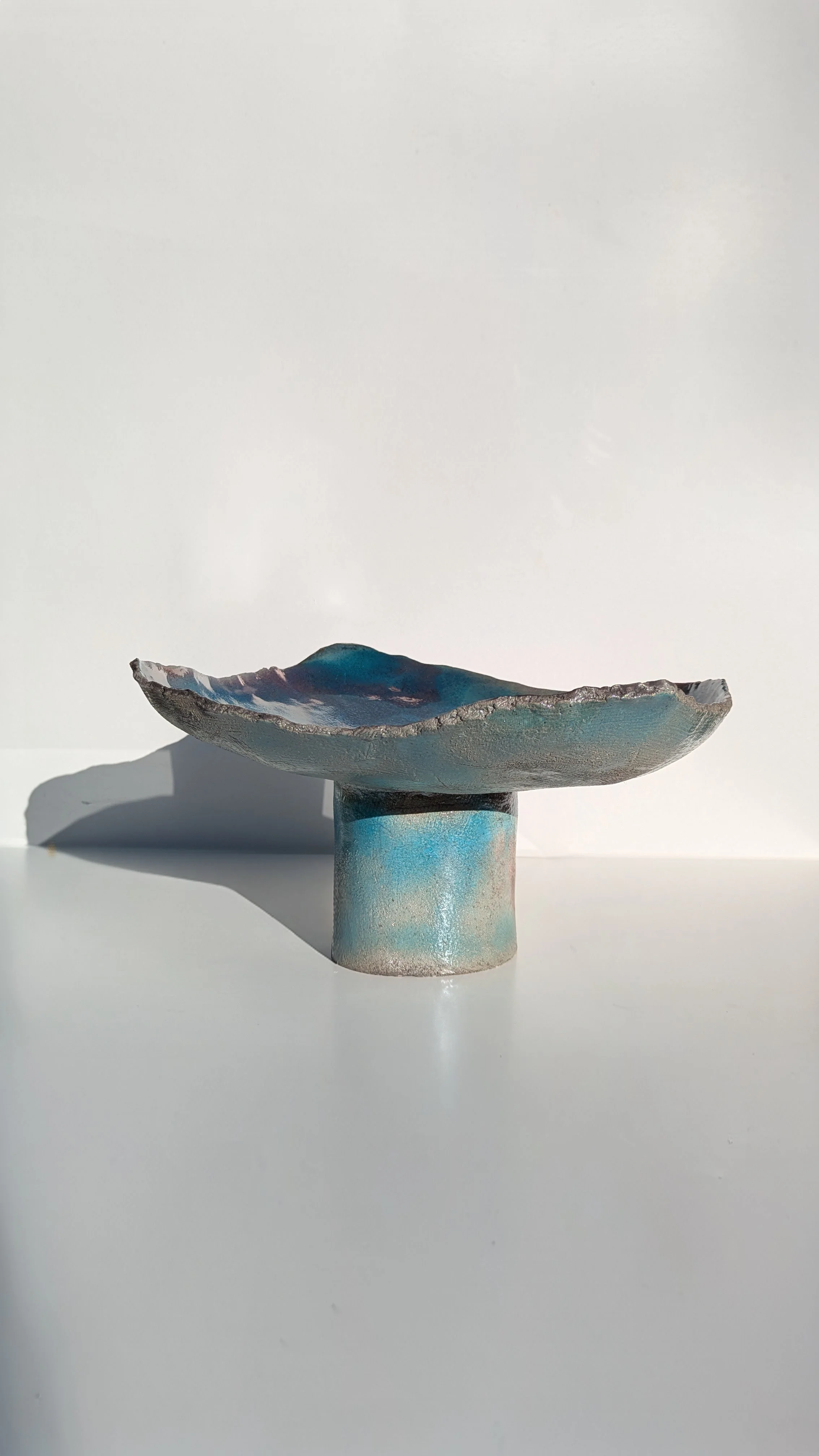 A sculptural ceramic bowl with a wide, irregular rim, placed on a cylindrical pedestal, against a plain white background. The bowl has a textured, glazed surface with shades of blue, gray, and hints of pink.