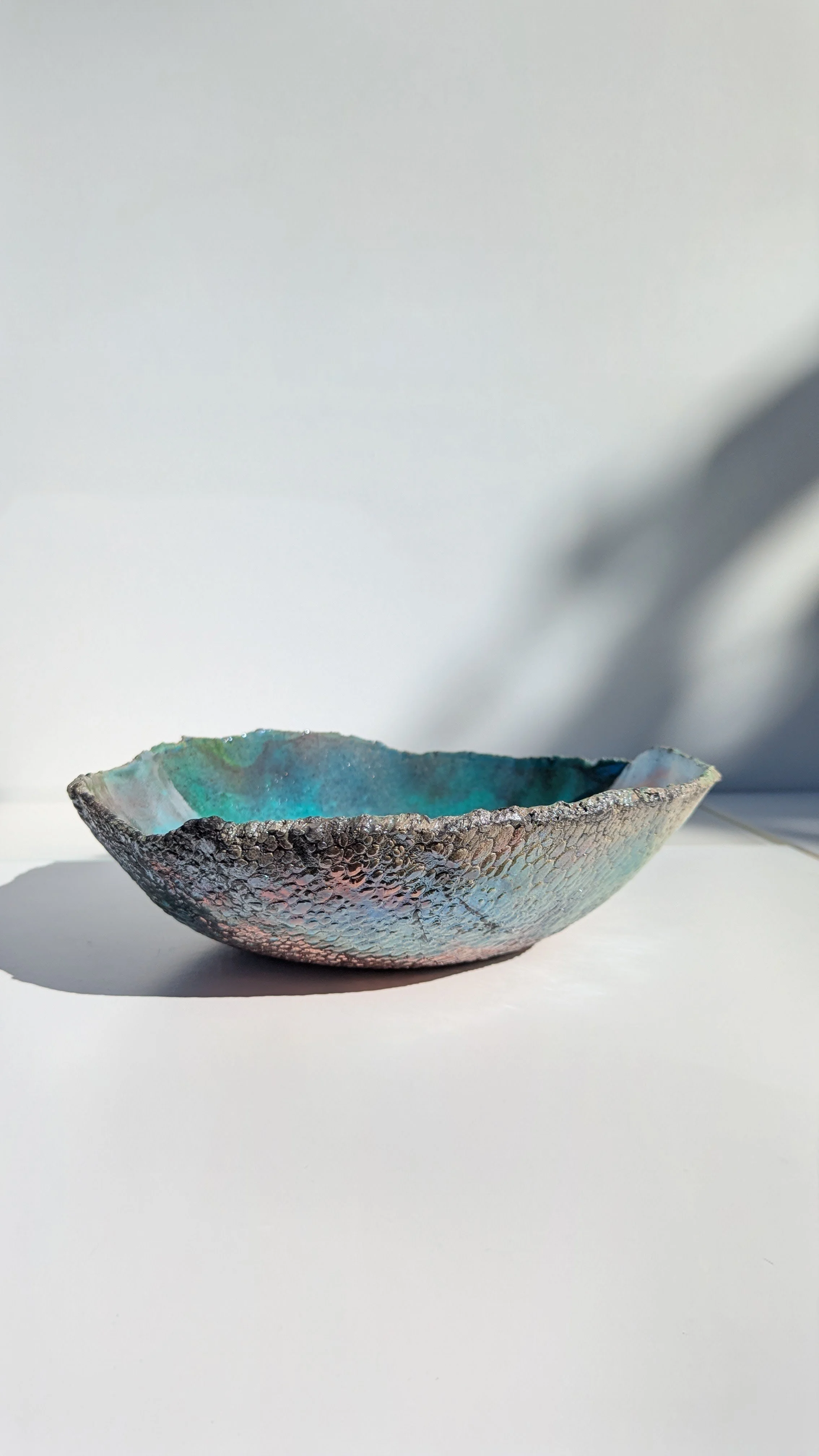 A textured metallic bowl with an iridescent interior, placed on a white surface with shadows cast on the background.