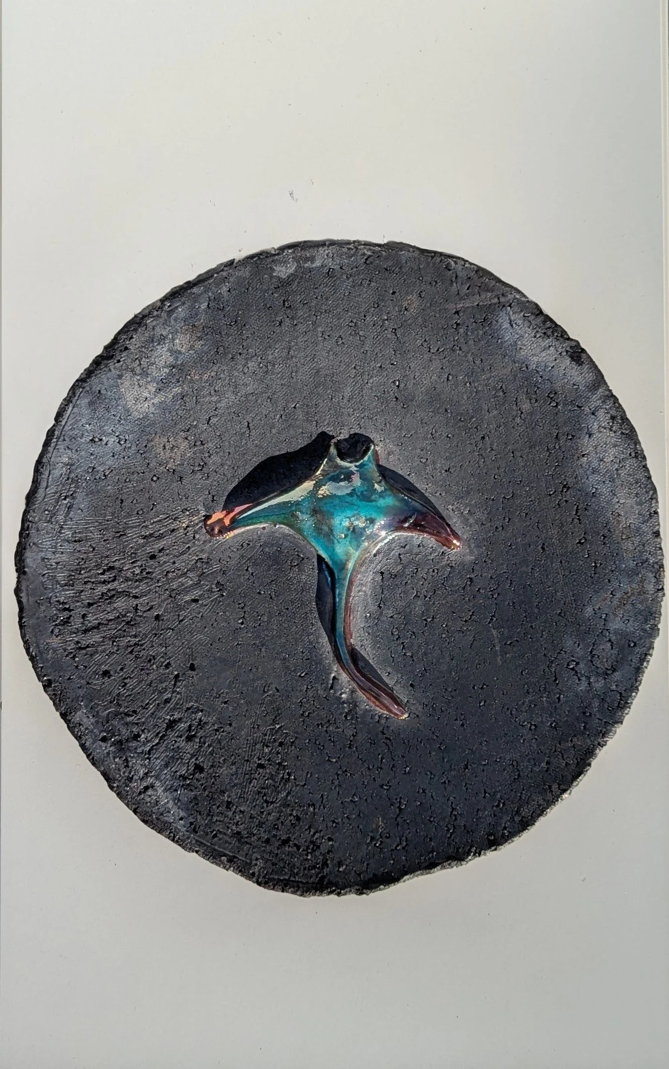 A black circular textured artwork with a small, shiny, multicolored fish sculpture at the center.