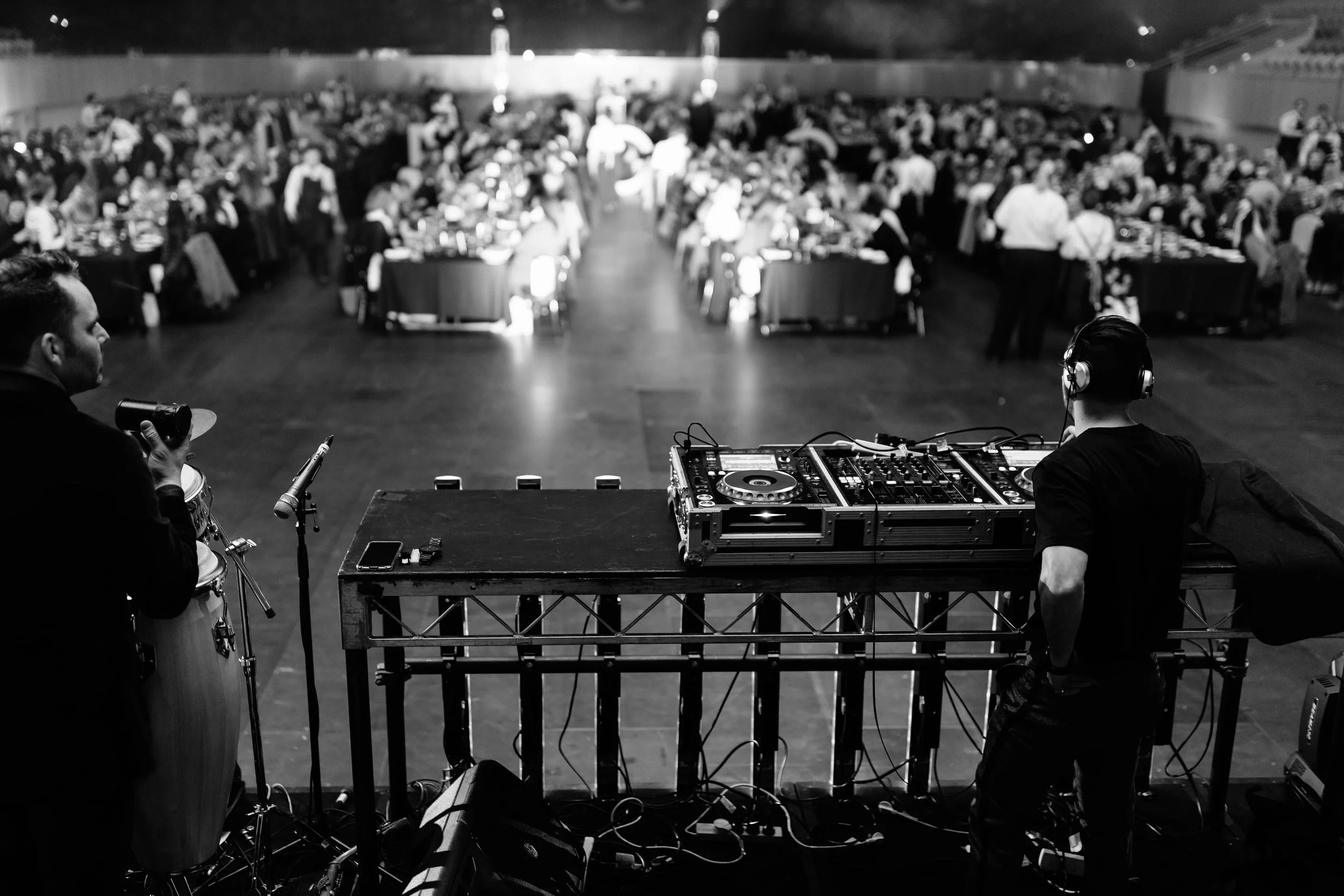 Black and white photo of a DJ mixing music on a stage at a large event, with a crowded dance floor and banquet tables in the background.