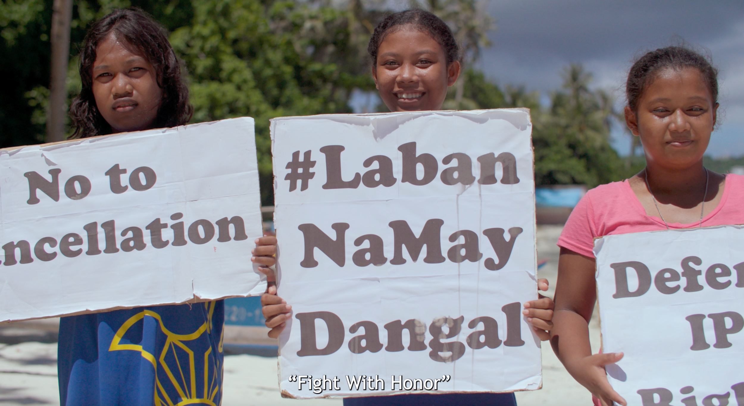 Three young women standing outdoors on a sunny day, holding signs protesting derogatory language and defending their honor, with trees and cloudy sky in the background.