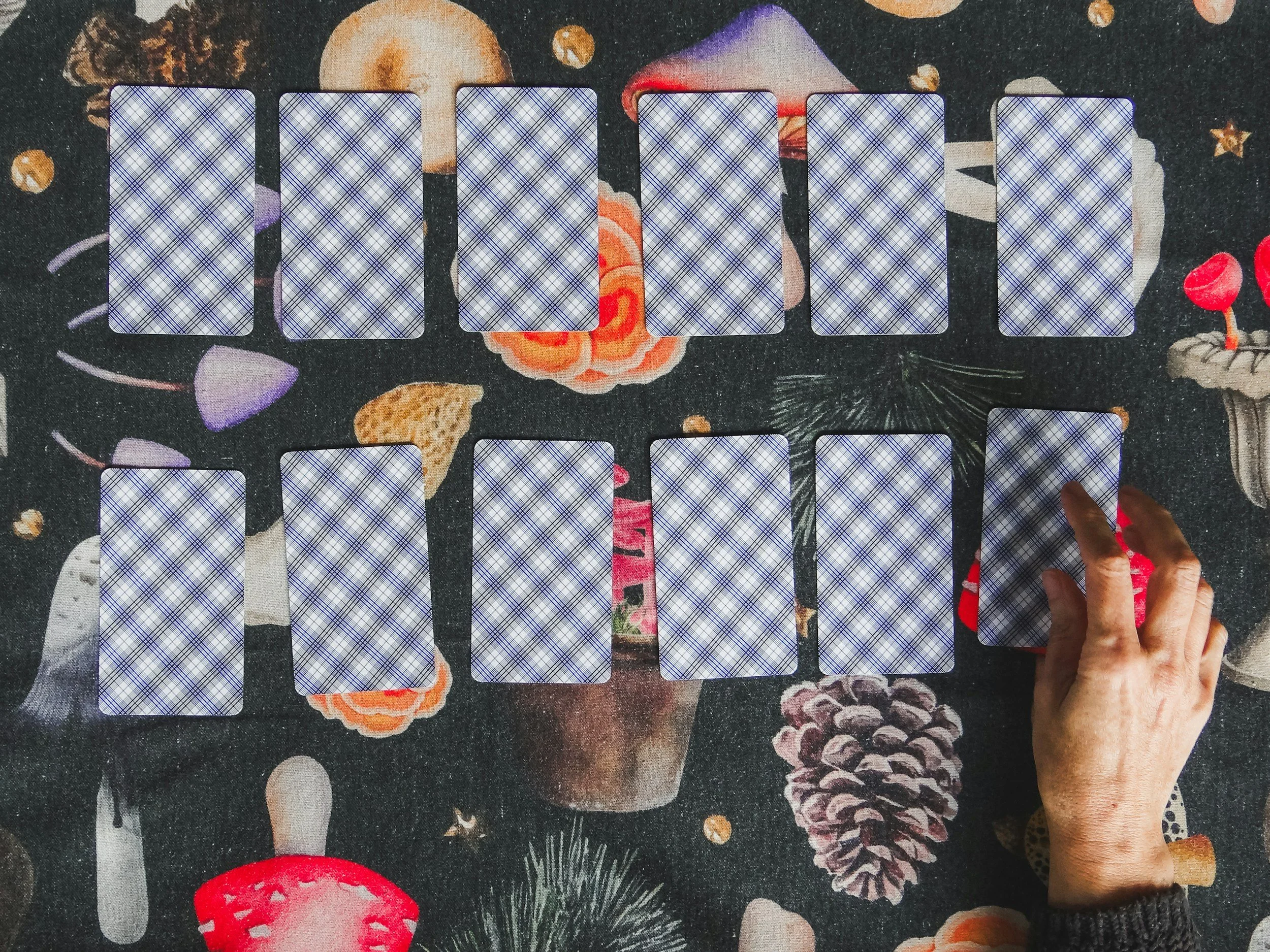 A person's hand reaches across a table to pick up a card from a set of tarot cards laid out in two rows on a dark, festive tablecloth decorated with pinecones, mushrooms, and holly.