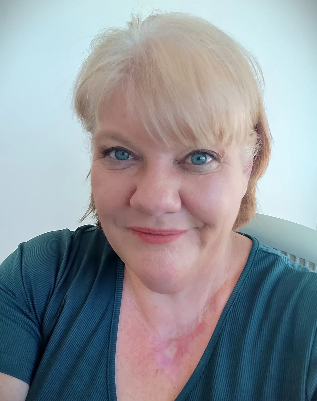 Close-up of smiling older woman with short blonde hair and blue eyes, wearing a teal shirt, against a light background.