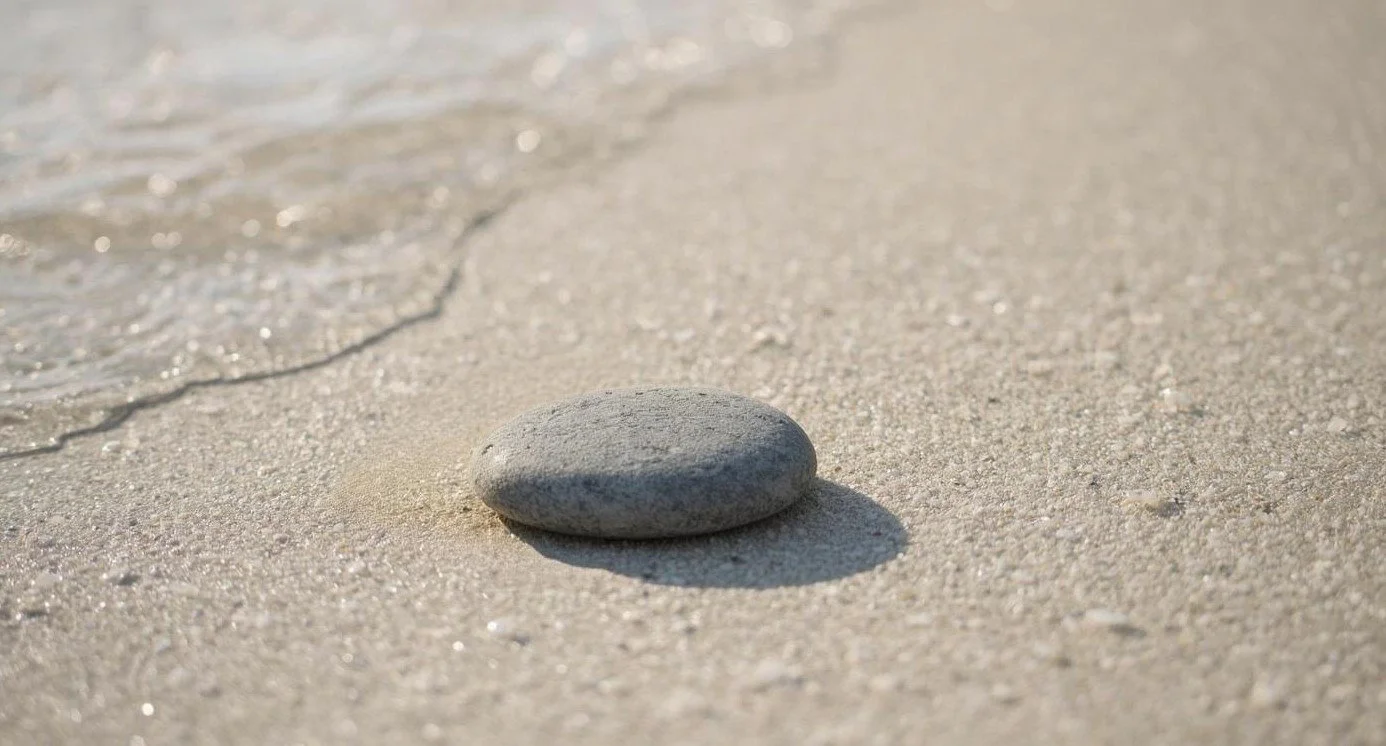 Smooth gray stone on sandy beach with ocean wave in the background.