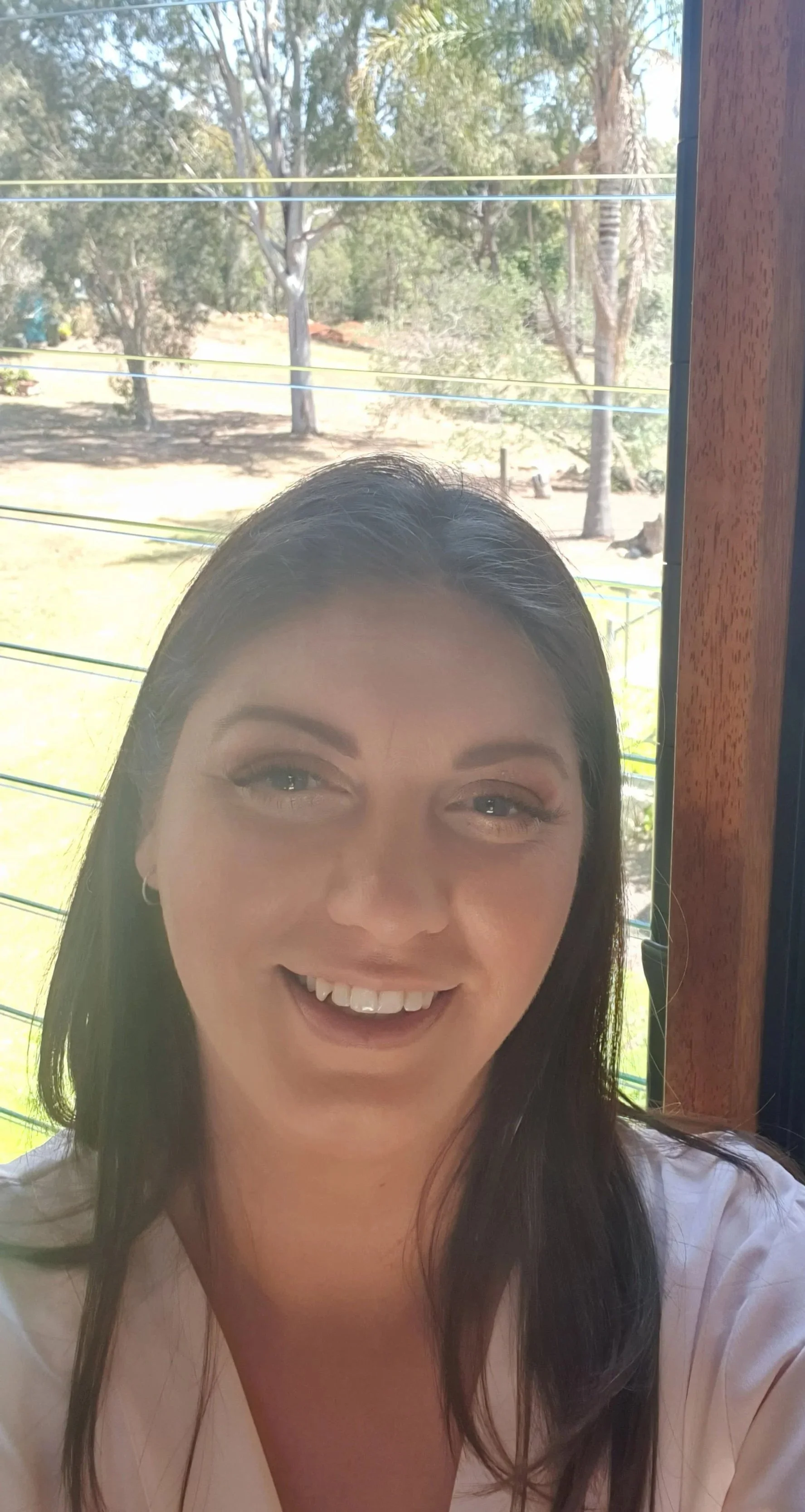 A woman with dark hair smiling in front of a window overlooking an outdoor area with trees and power lines.
