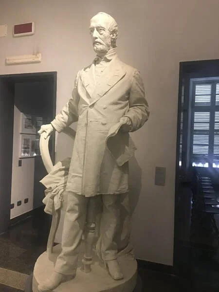 White marble statue of Giuseppe Mazzini standing indoors, holding a rolled document in his right hand and resting his left hand on a decorative chair.