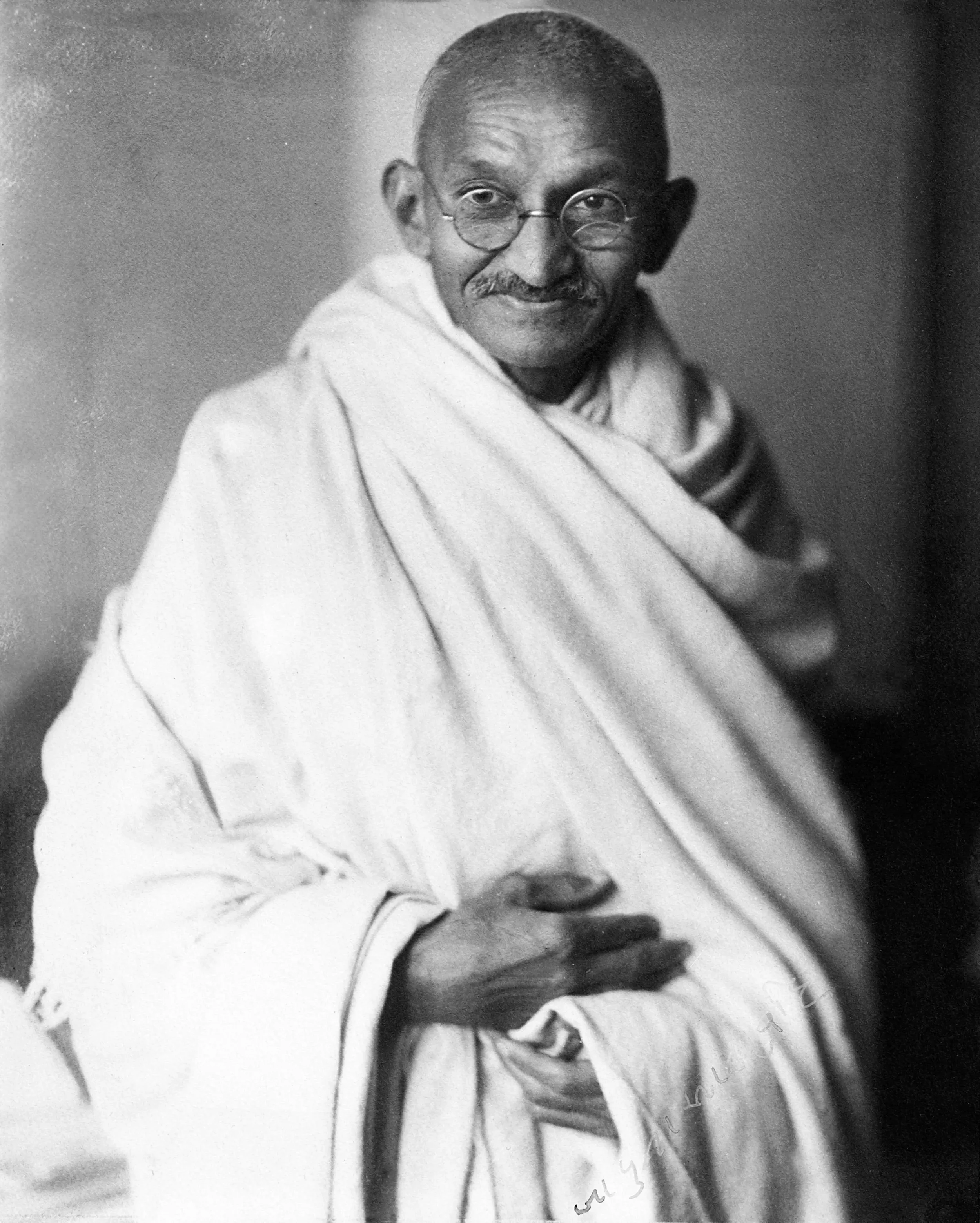 A black and white photograph of Mahatma Gandhi, an Indian leader, wearing round glasses and traditional Indian attire, smiling gently and holding his hands together.