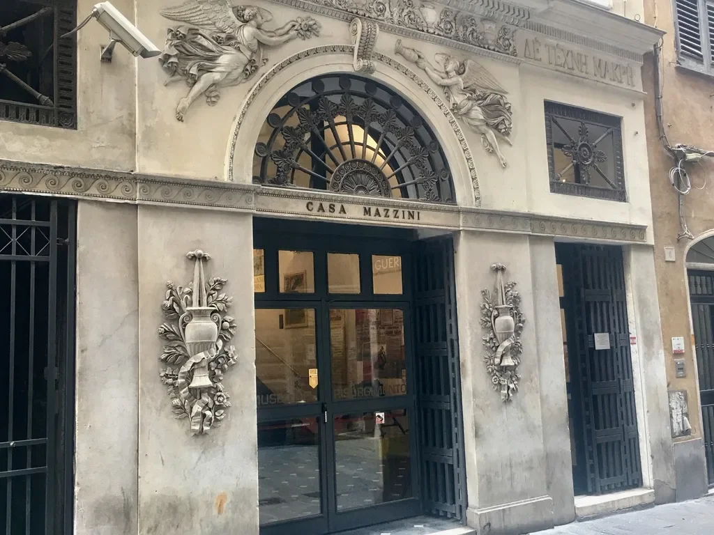 The exterior of Casa Mazzini, a building with ornate architectural details, sculptures, and decorative elements on the facade. The entrance is framed by black gates and a semi-circular window above.