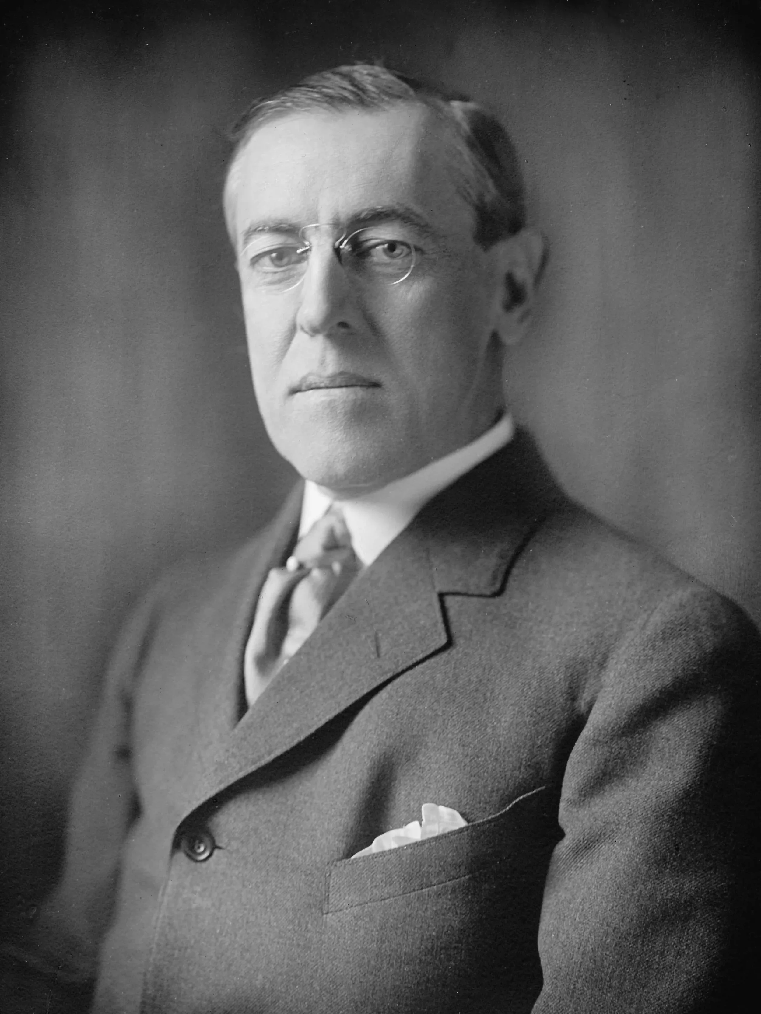 Black and white portrait of a Woodrow Wilson wearing a suit, tie, and glasses, with short hair, looking serious.