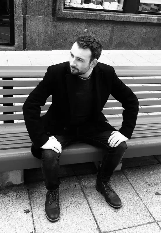 A man with short dark hair and a beard sitting on a wooden park bench outdoors, dressed in black jacket, black shirt, black pants, and black boots, looking to his left.