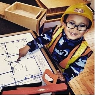 Young boy wearing safety helmet and glasses, smiling, working on a building floor plan at a desk with a ruler, in a woodworking or classroom setting.