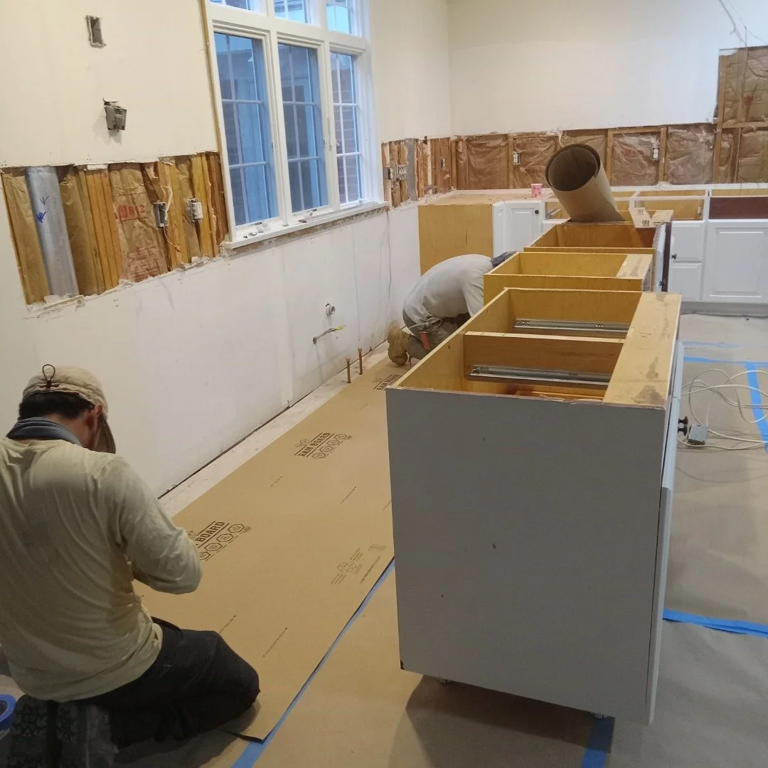 Two workers installing kitchen cabinets in a wall with exposed wiring and pipes, with a large window on the back wall and construction materials on the floor.