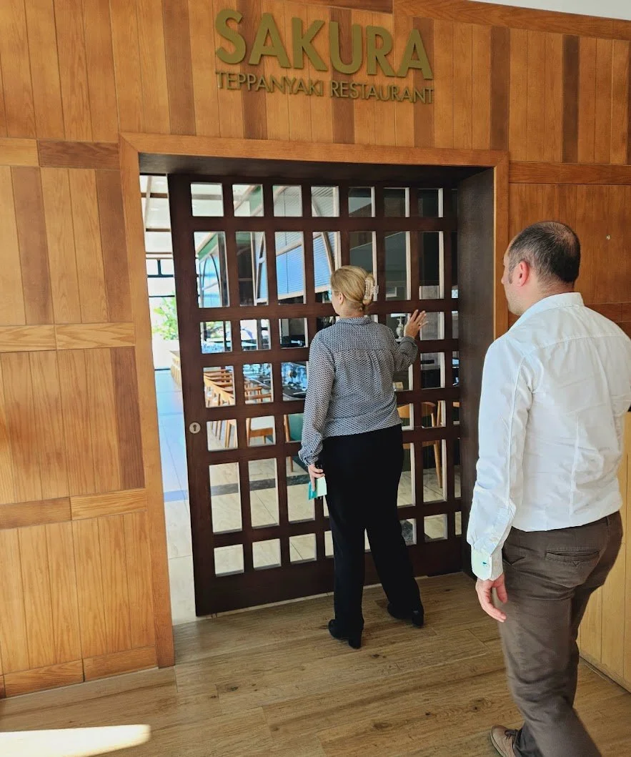 Two people standing near the entrance of Sakura Teppanyaki Restaurant with a wooden lattice door and a wooden wall. One person is opening the door while the other is waiting.