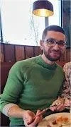 A man with glasses and a beard wearing a green sweater, sitting at a table in a restaurant.