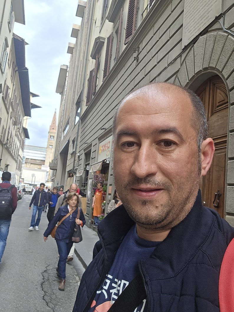 A man taking a selfie on a crowded European city street with historic buildings and a clock tower in the background.