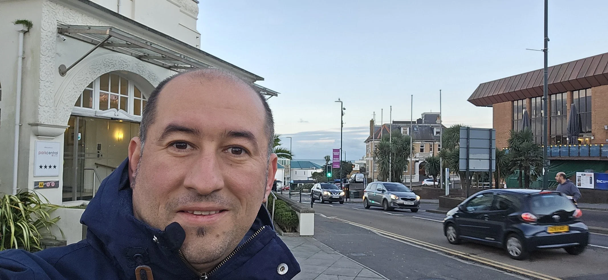 A man taking a selfie on a city street with cars passing by, palm trees, and buildings under a blue sky.