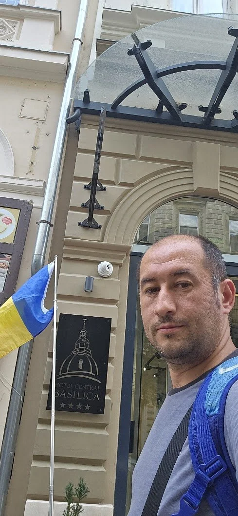 Selfie of a man taking a photo outside the Hotel Central Basilica, with a gray building, a Ukrainian flag, and a sign featuring a city icon in the background.