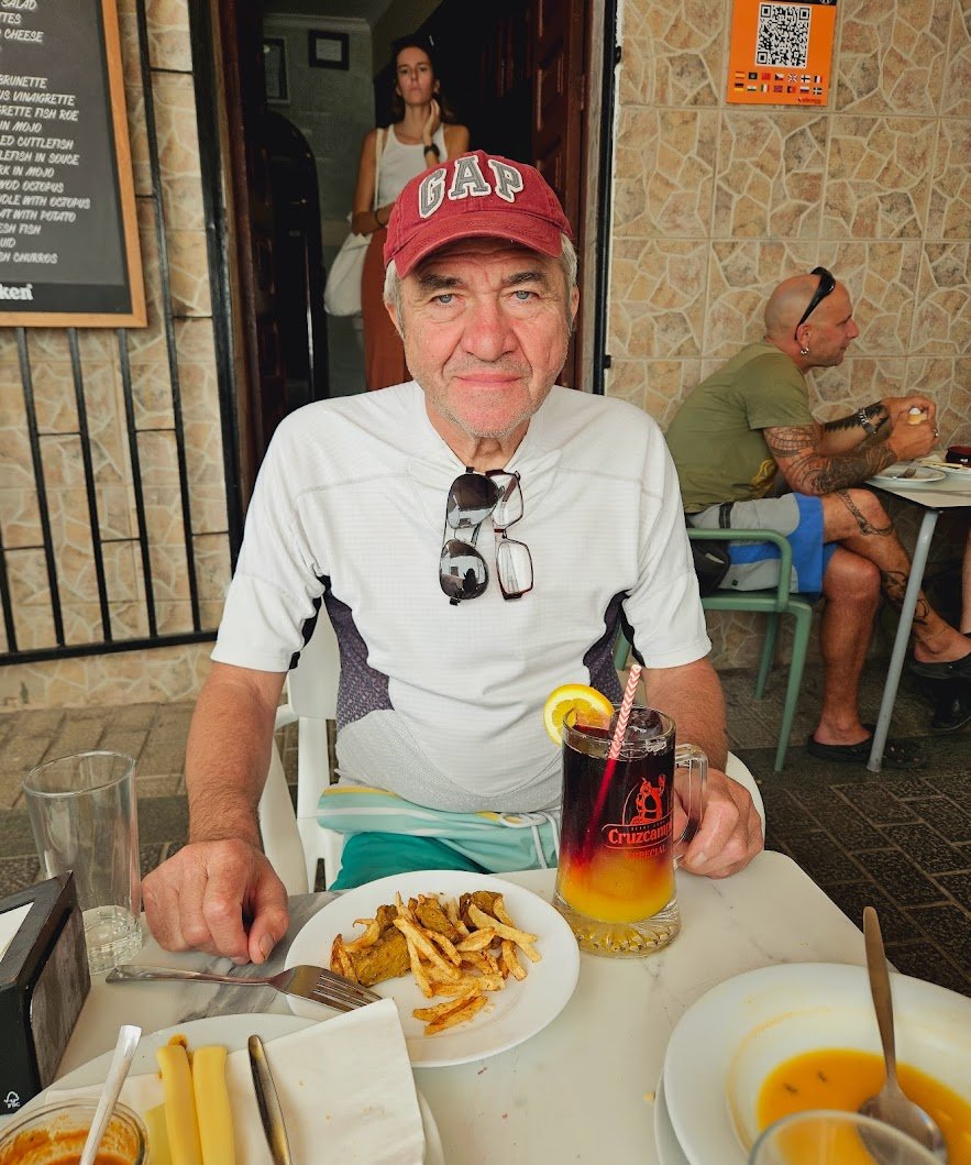 Older man wearing a white shirt, red GAP cap, and sunglasses hanging on his shirt, sitting at a table with a plate of fries, a large drink with lemon and a straw. Behind him, a bald man with tattoos in a green shirt and blue shorts is seated at anoth