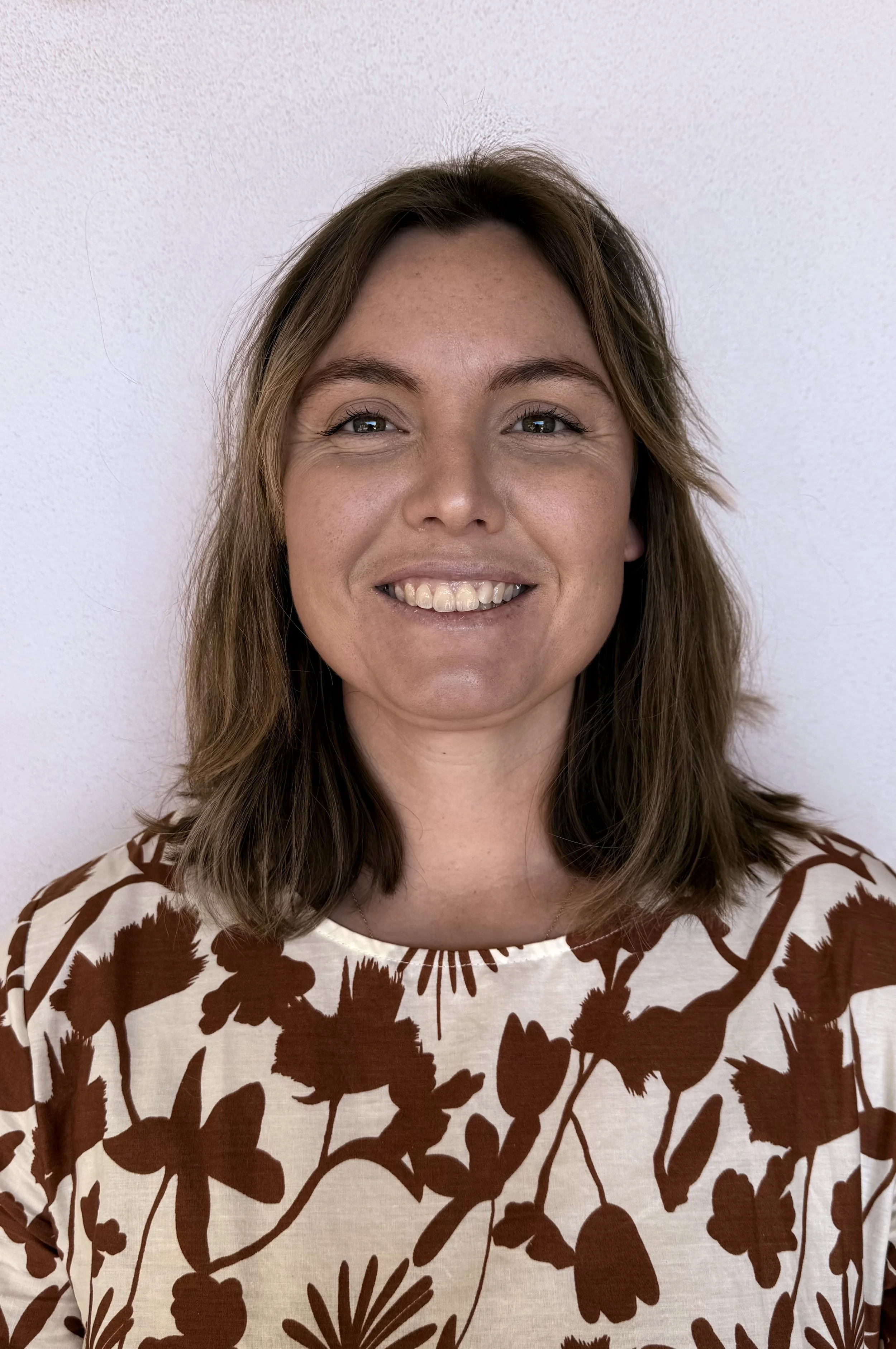 A woman with shoulder-length brown hair smiling at the camera, wearing a cream-colored shirt with brown floral patterns, in front of a plain light-colored wall.