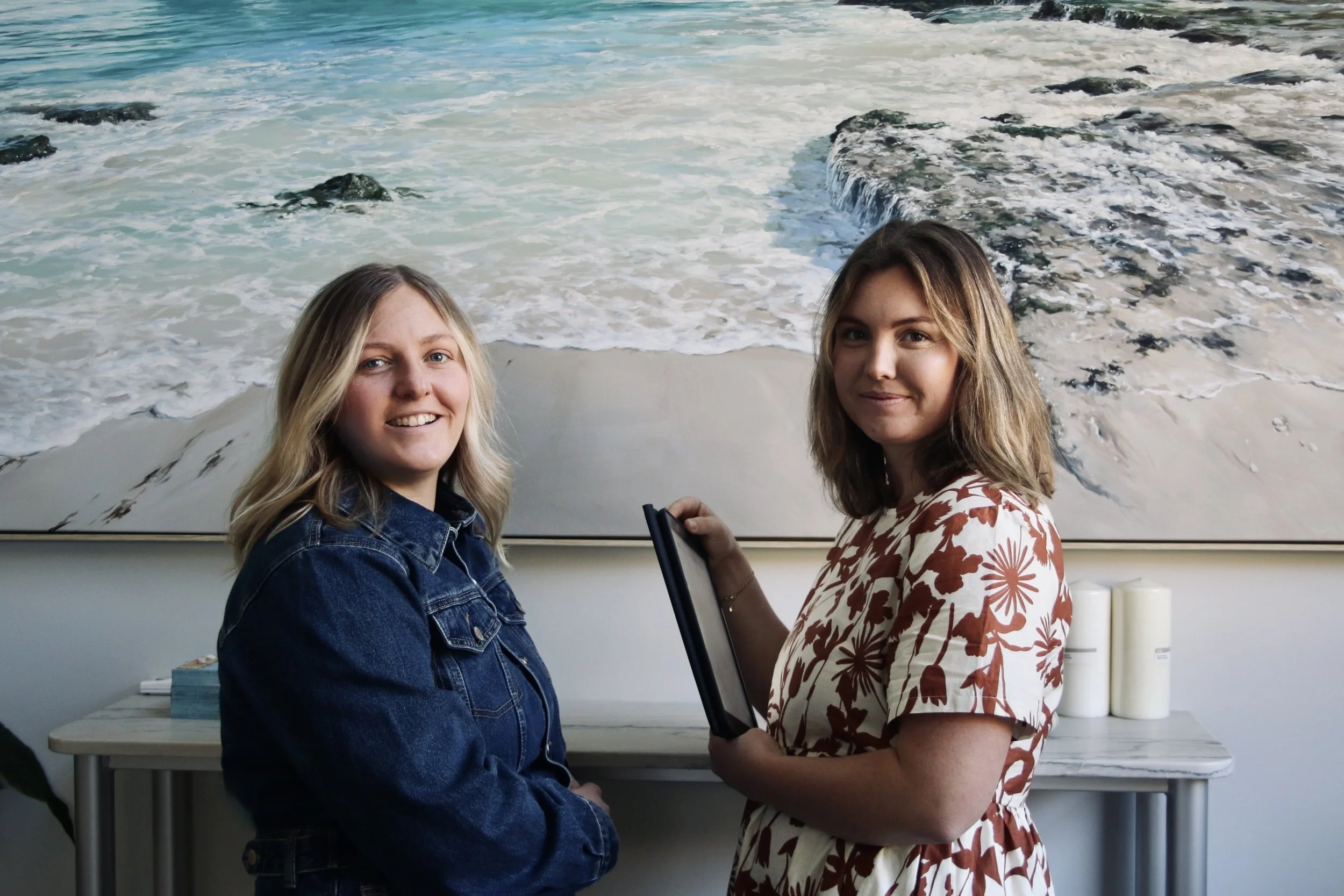 Two women standing indoors in front of a large painting of a beach scene. One woman is wearing a denim jacket and the other is wearing a floral dress and holding a folder.
