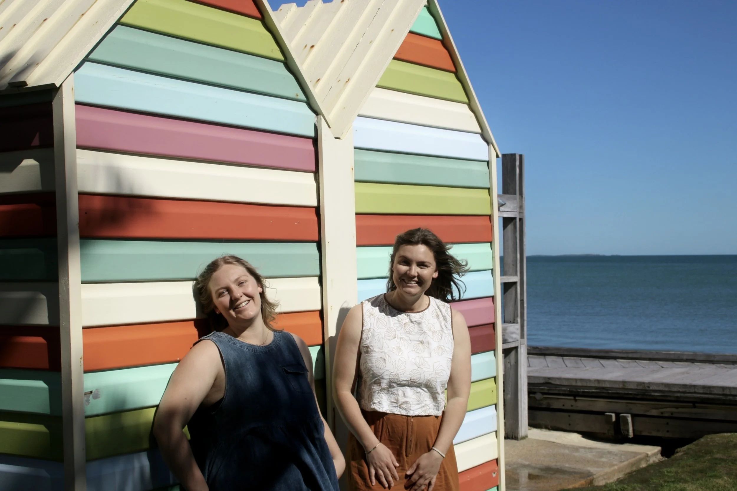 Two women smiling and standing outside near a colorful rainbow-striped beach hut with the ocean and blue sky in the background.