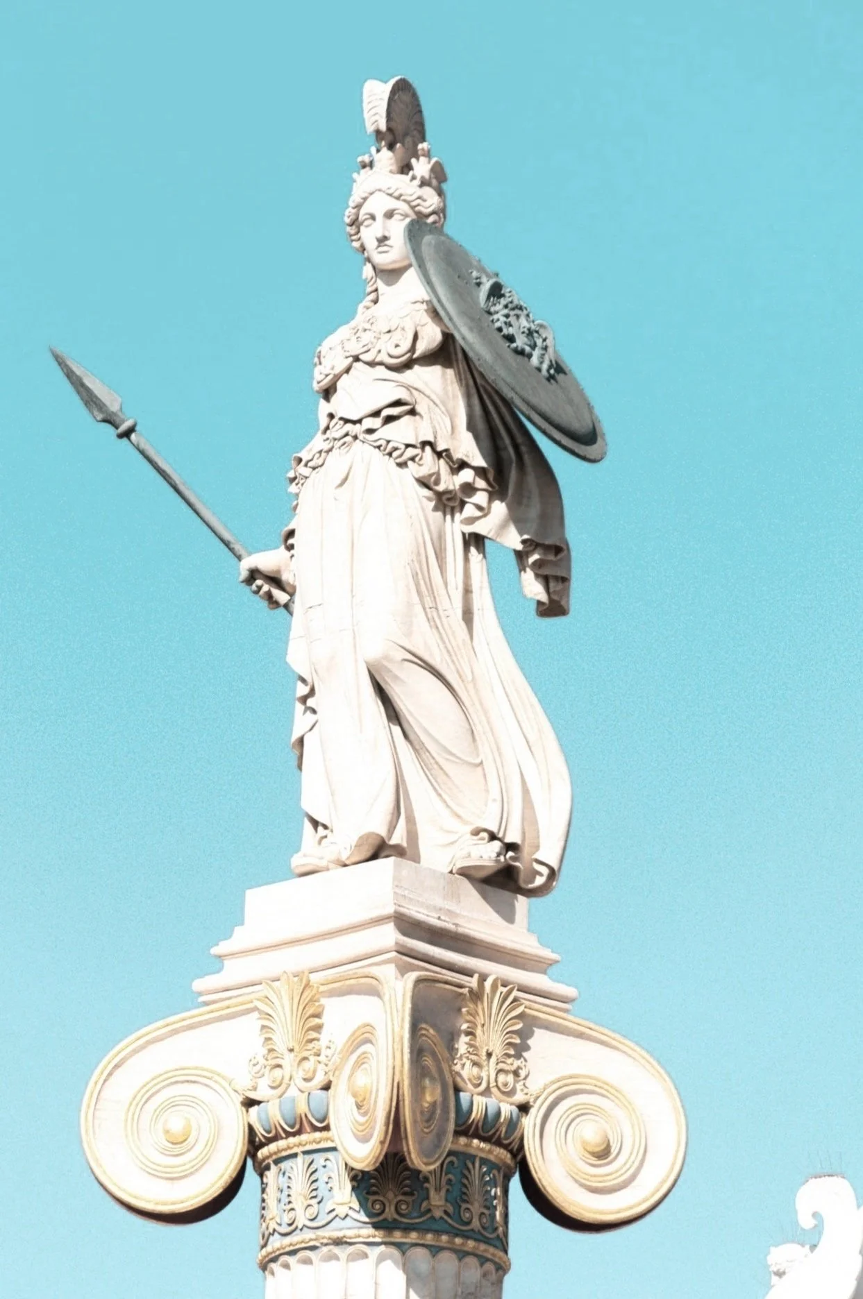 Statue of a classical female warrior holding a spear in her right hand and a shield in her left, standing on a decorated column against a bright blue sky.