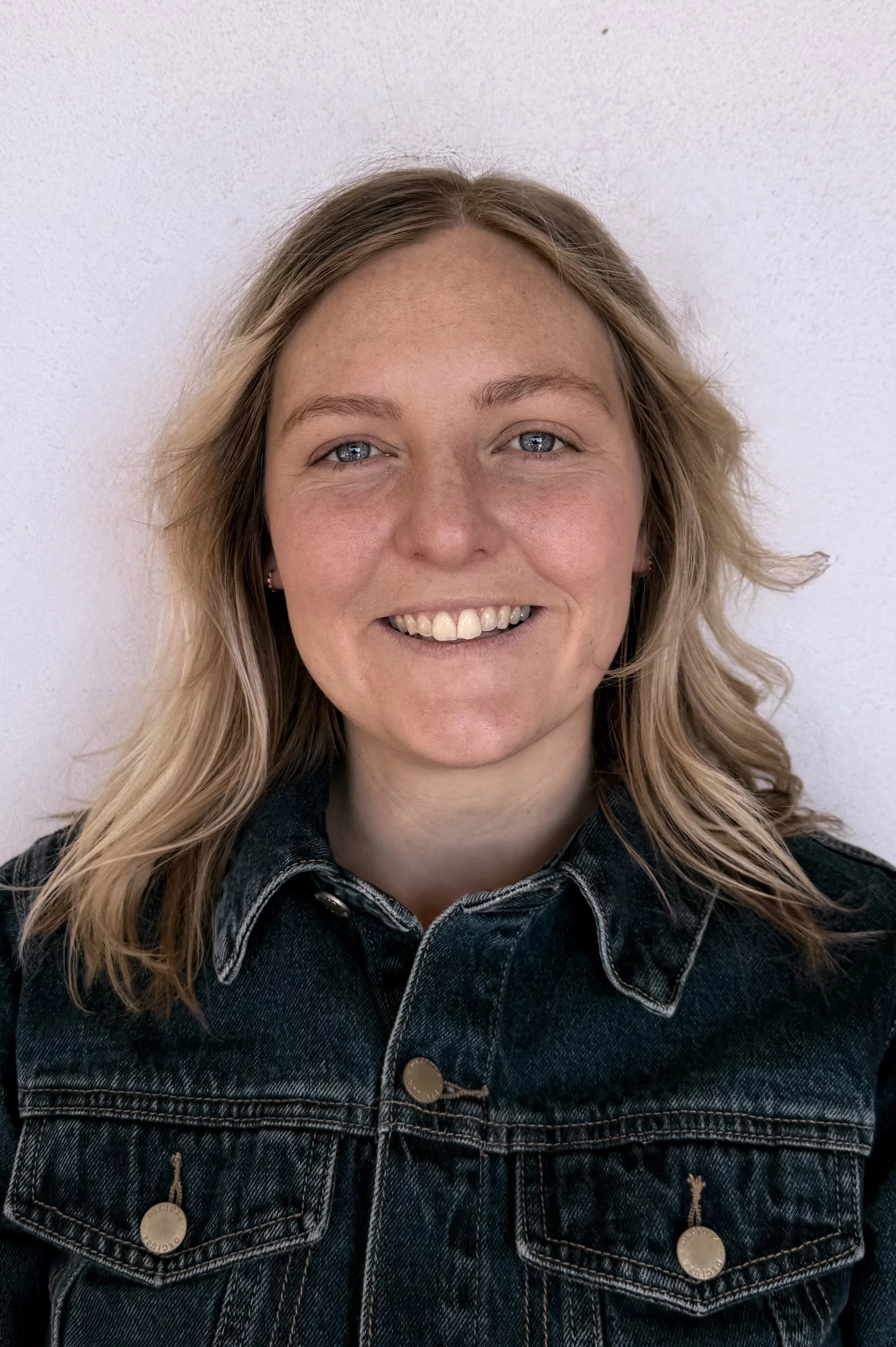 A woman with blonde hair and blue eyes smiling, wearing a denim jacket, standing against a plain white wall.