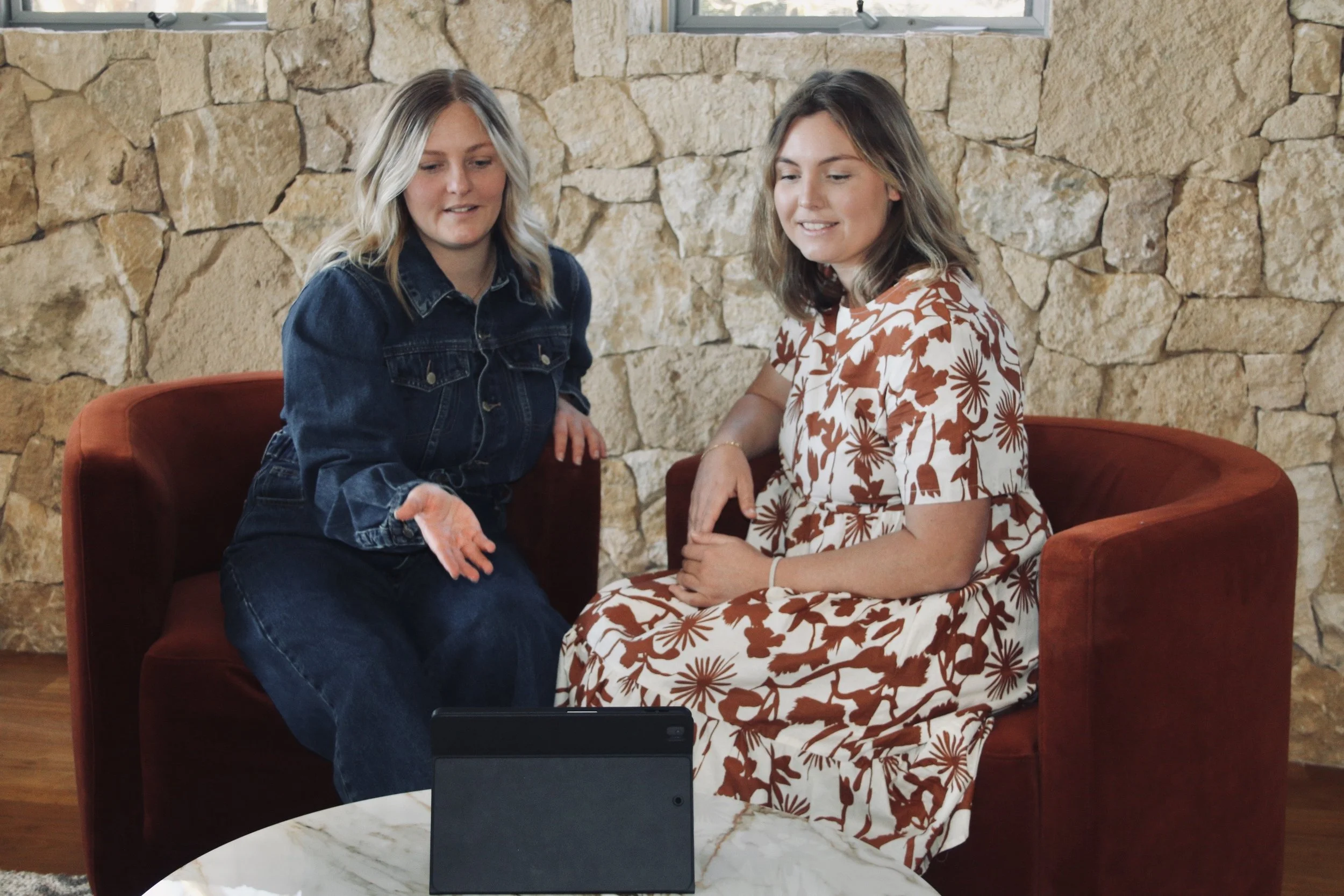 Two women sitting on a red couch in front of a stone wall, engaged in a conversation with a tablet on a white marble table in front of them.